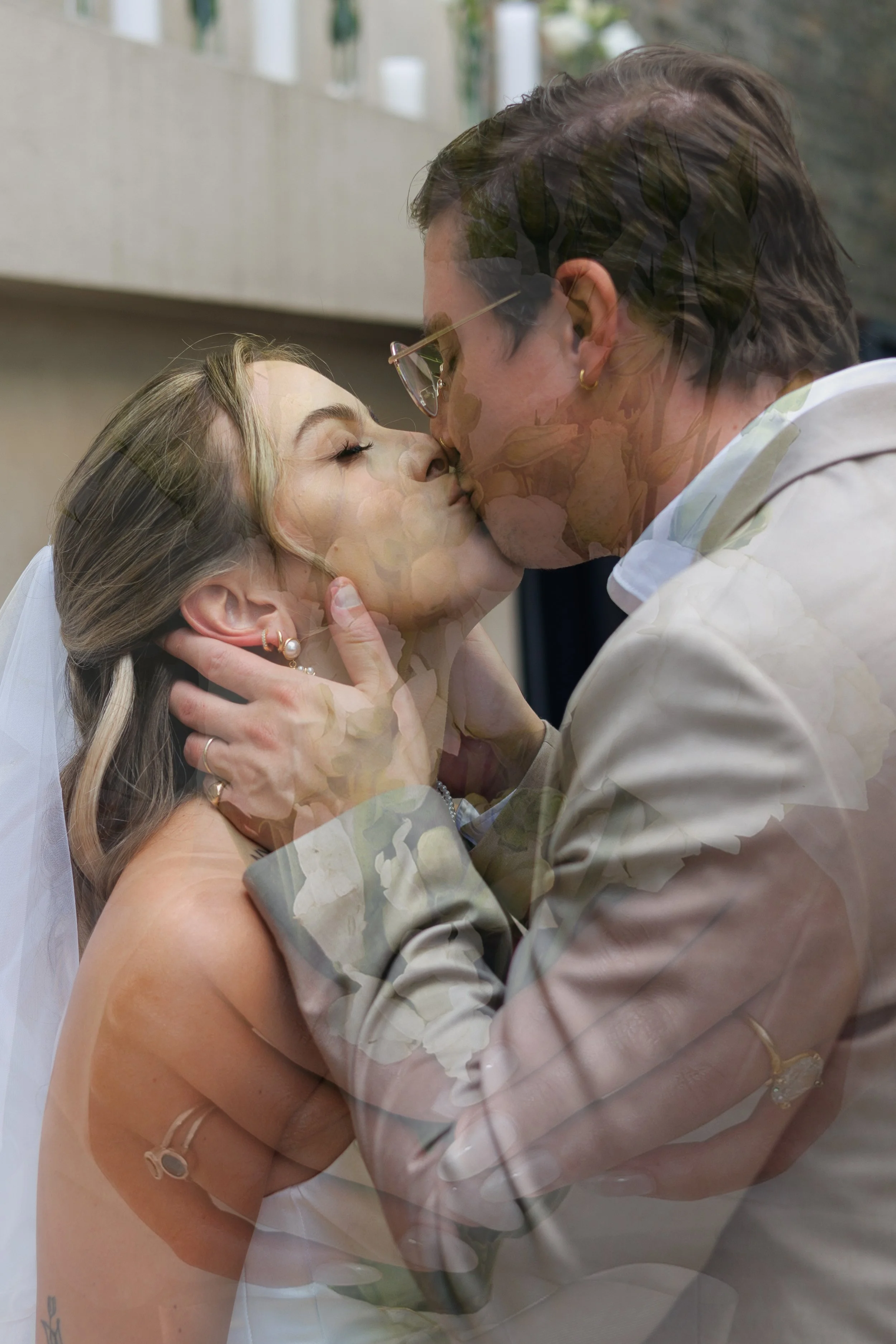 A man and woman sharing a kiss at a wedding, with the woman wearing a wedding dress and veil, and the man wearing glasses and a beige suit.