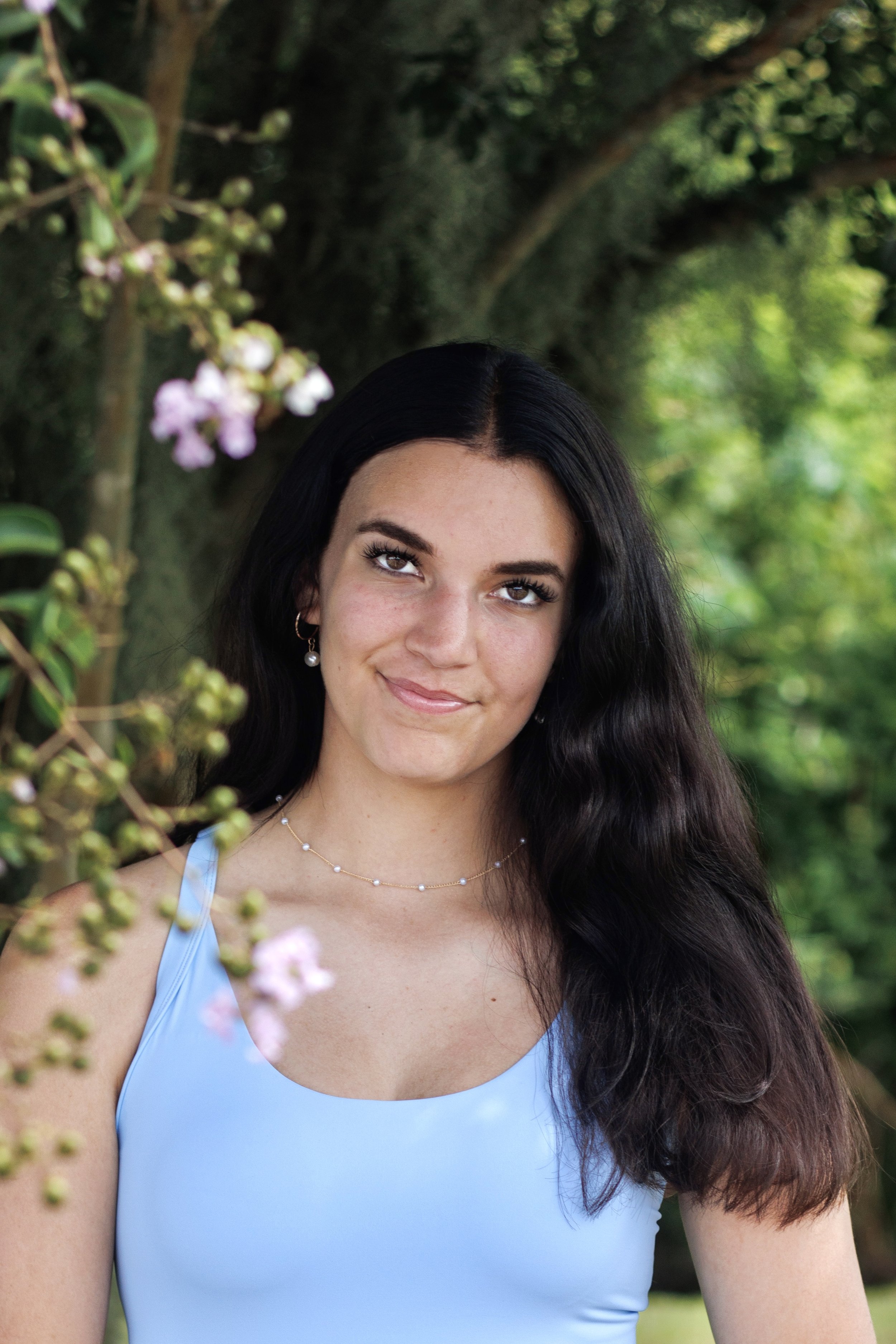 Woman in a blue tank top standing outdoors, surrounded by greenery and pink flowers.