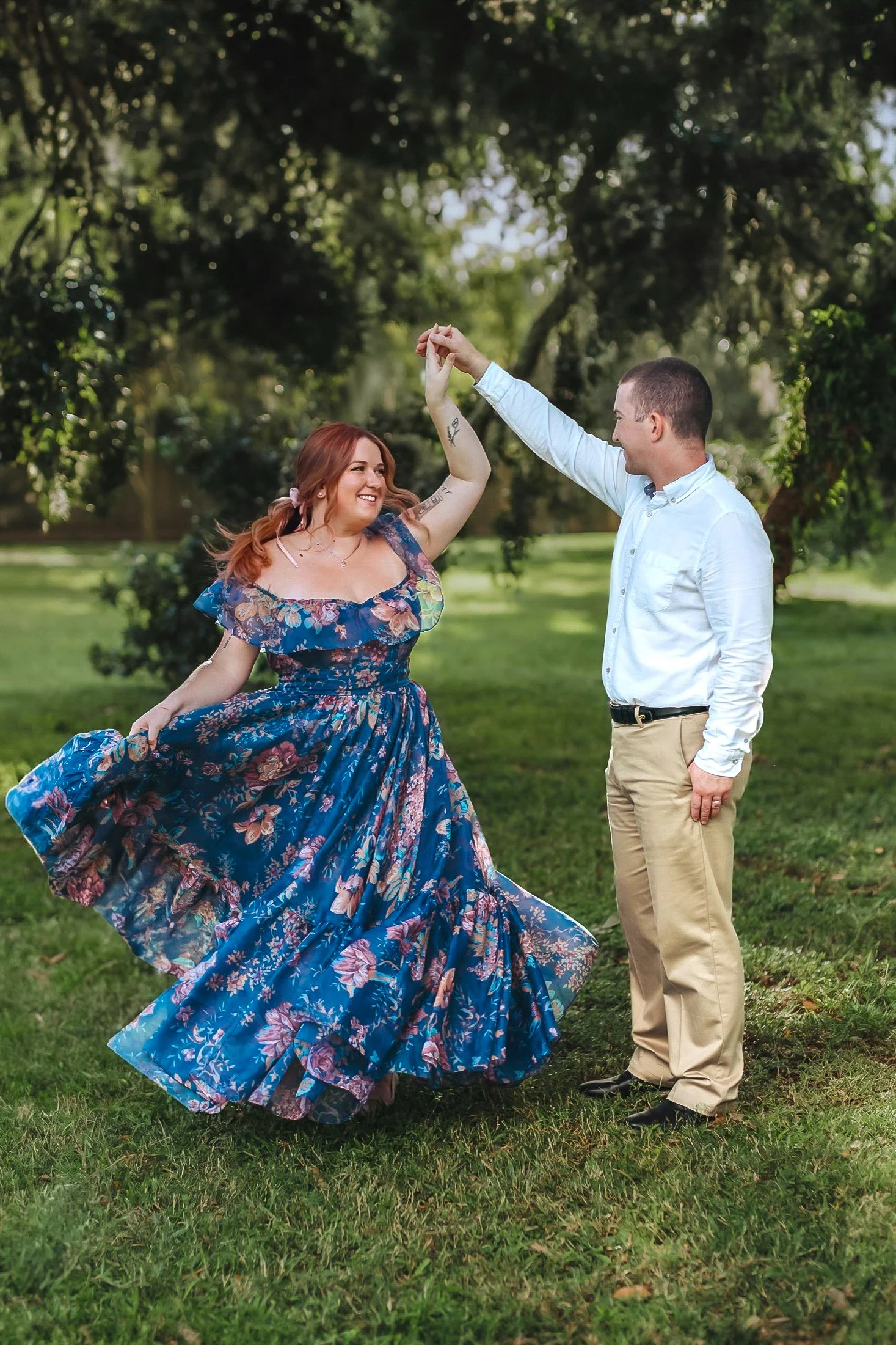 A couple dancing outdoors in a park, with the woman in a flowing, floral blue dress and the man in a white shirt and beige pants, holding hands and smiling.