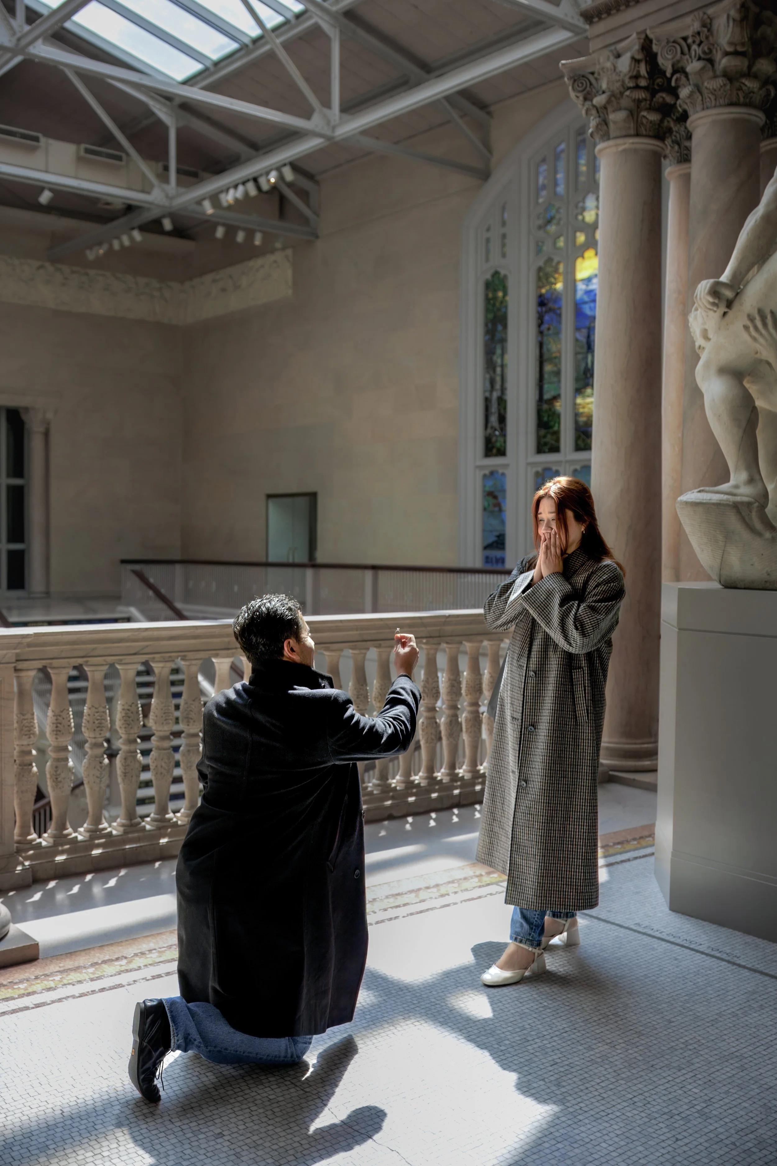 A man proposing marriage to a woman inside a church. The man is kneeling with a ring in his hand, and the woman is standing with her hands covering her mouth, appearing surprised. Sunlight filters through stained glass windows, illuminating the scene.