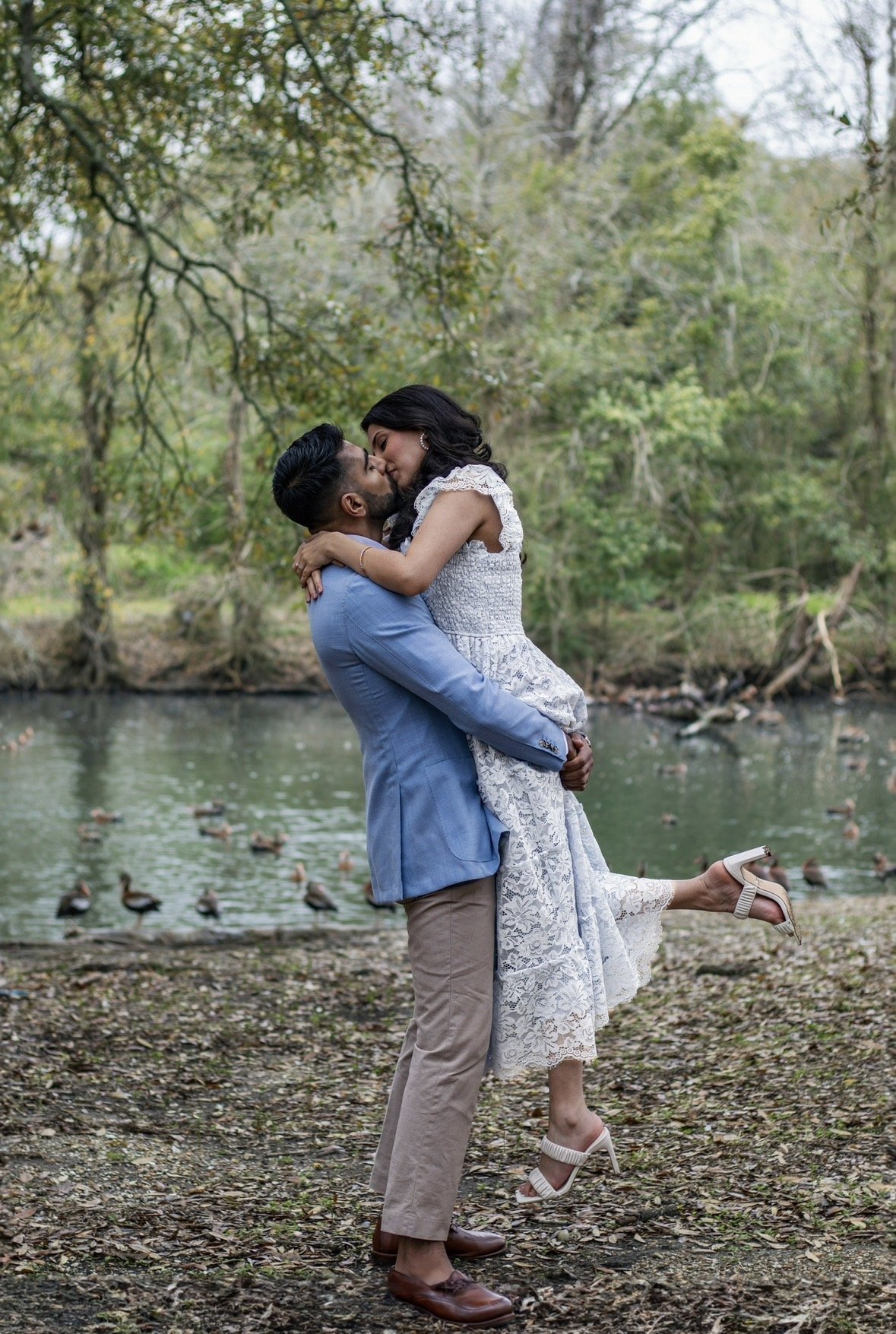 Chicago wedding photographer captures a couple kissing by a pond in a wooded area, with ducks swimming in the back. The man is wearing a blue blazer and khakis, and the woman is in a white lace dress and heels, with her legs lifted off the ground.