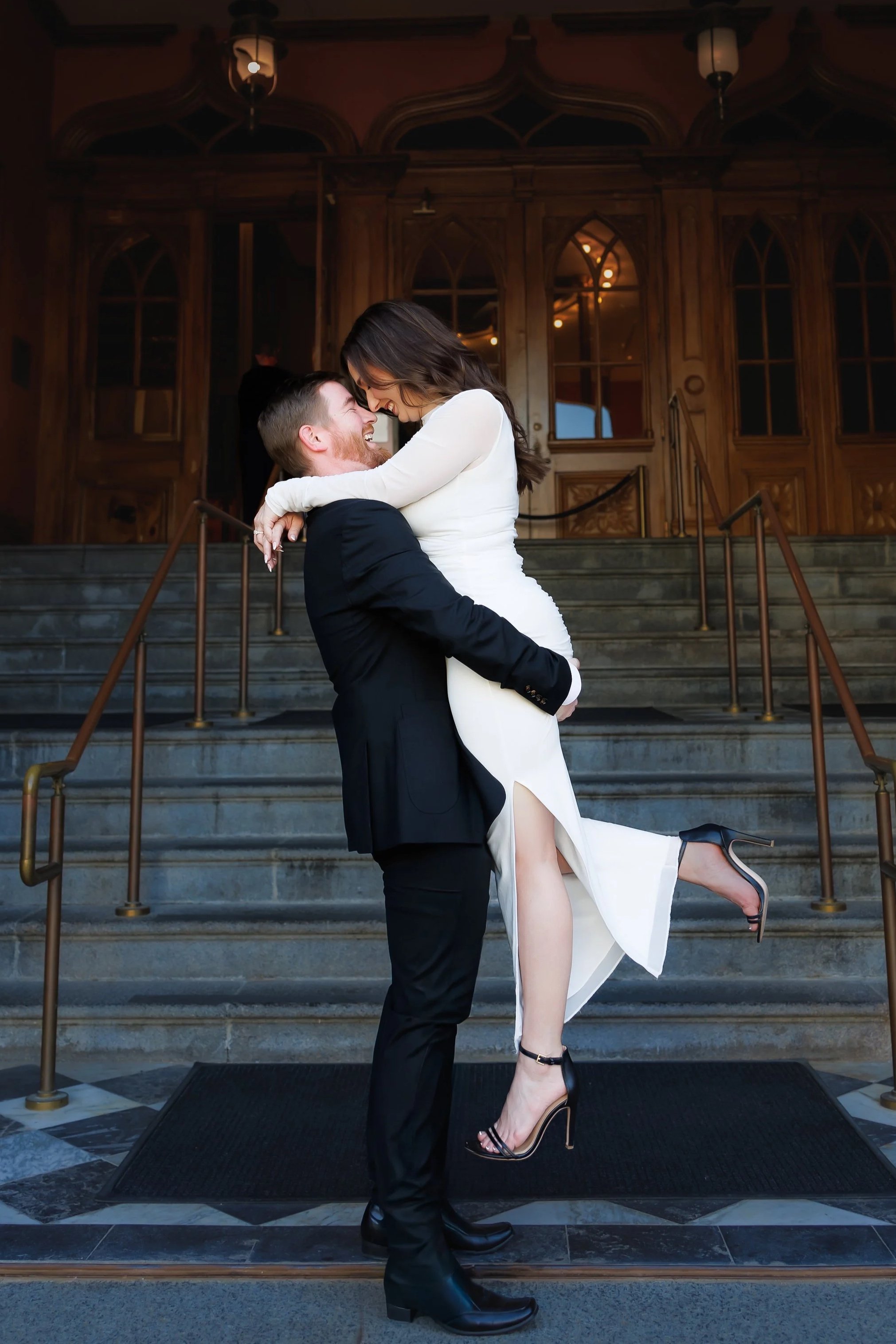 A couple walking hand in hand on a sidewalk, surrounded by columns, with the woman in a white dress and the man in a patterned shirt.