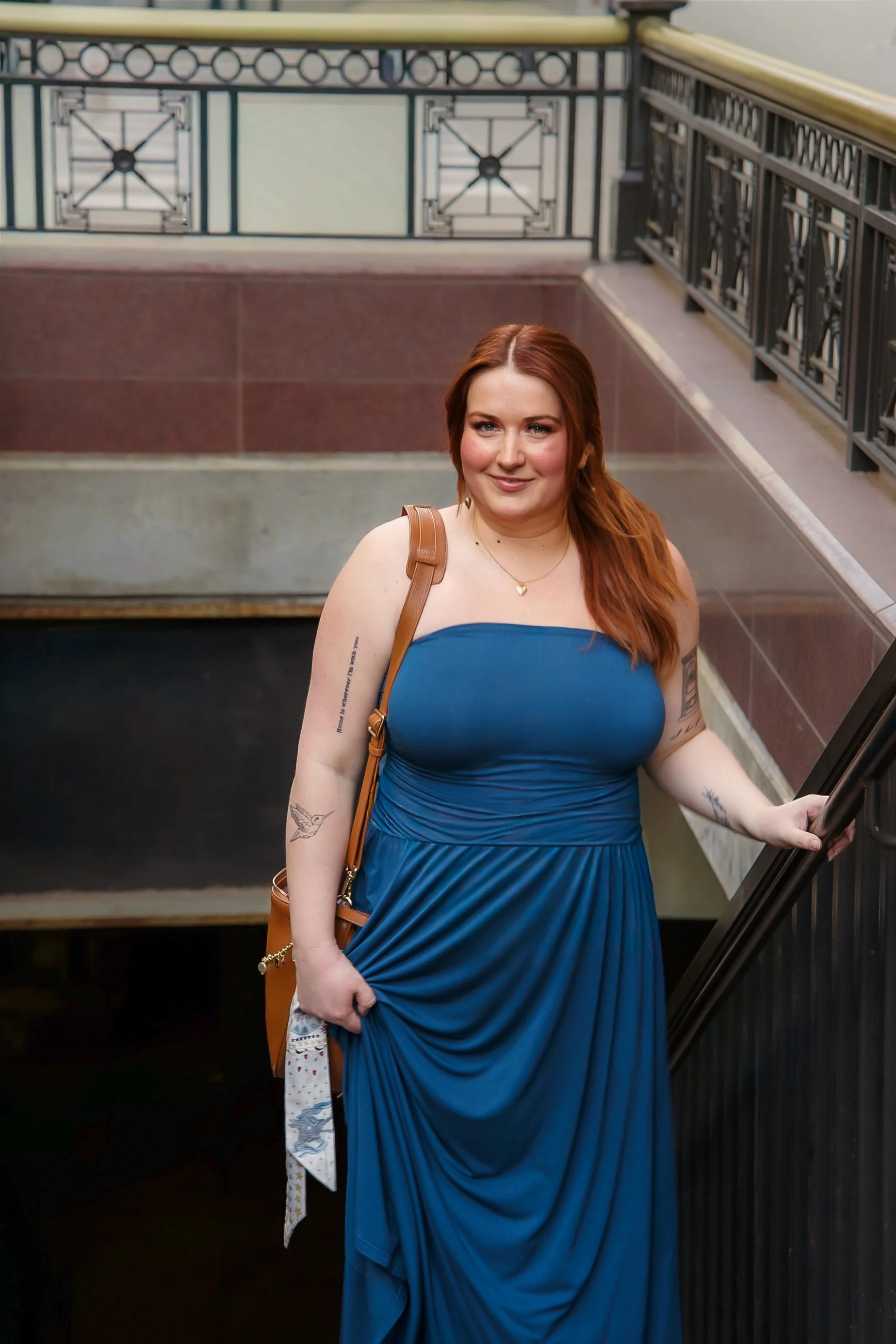 A woman with red hair in a blue dress standing on a staircase, holding the railing with her right hand, smiling.