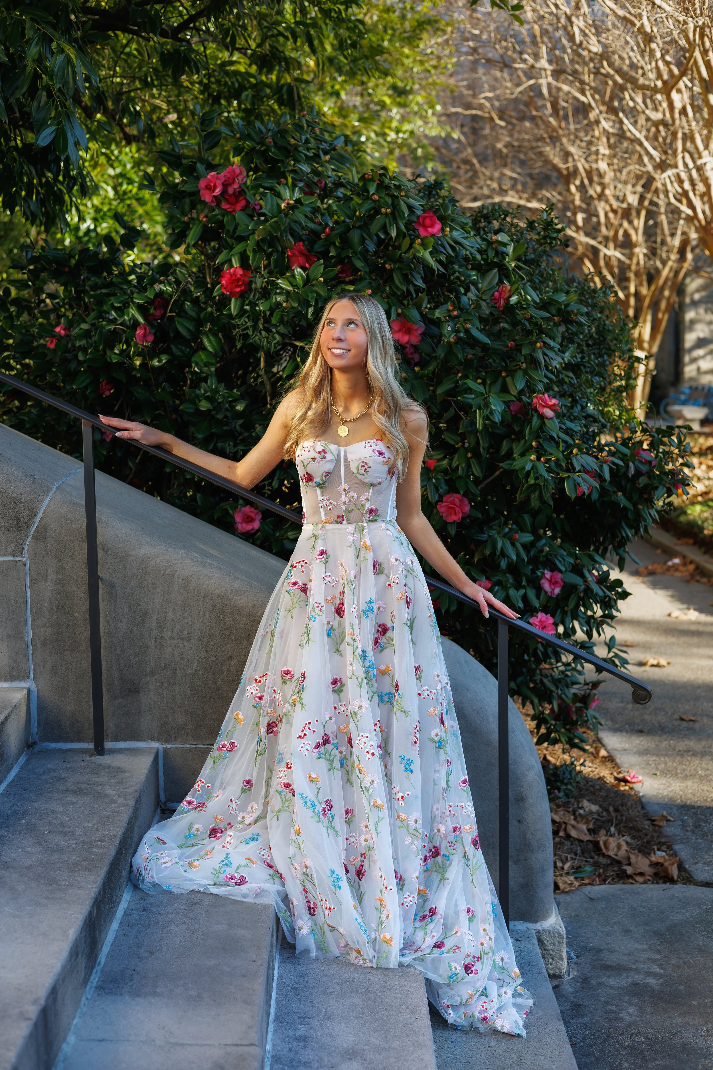 Woman in floral gown standing on steps near blooming bushes