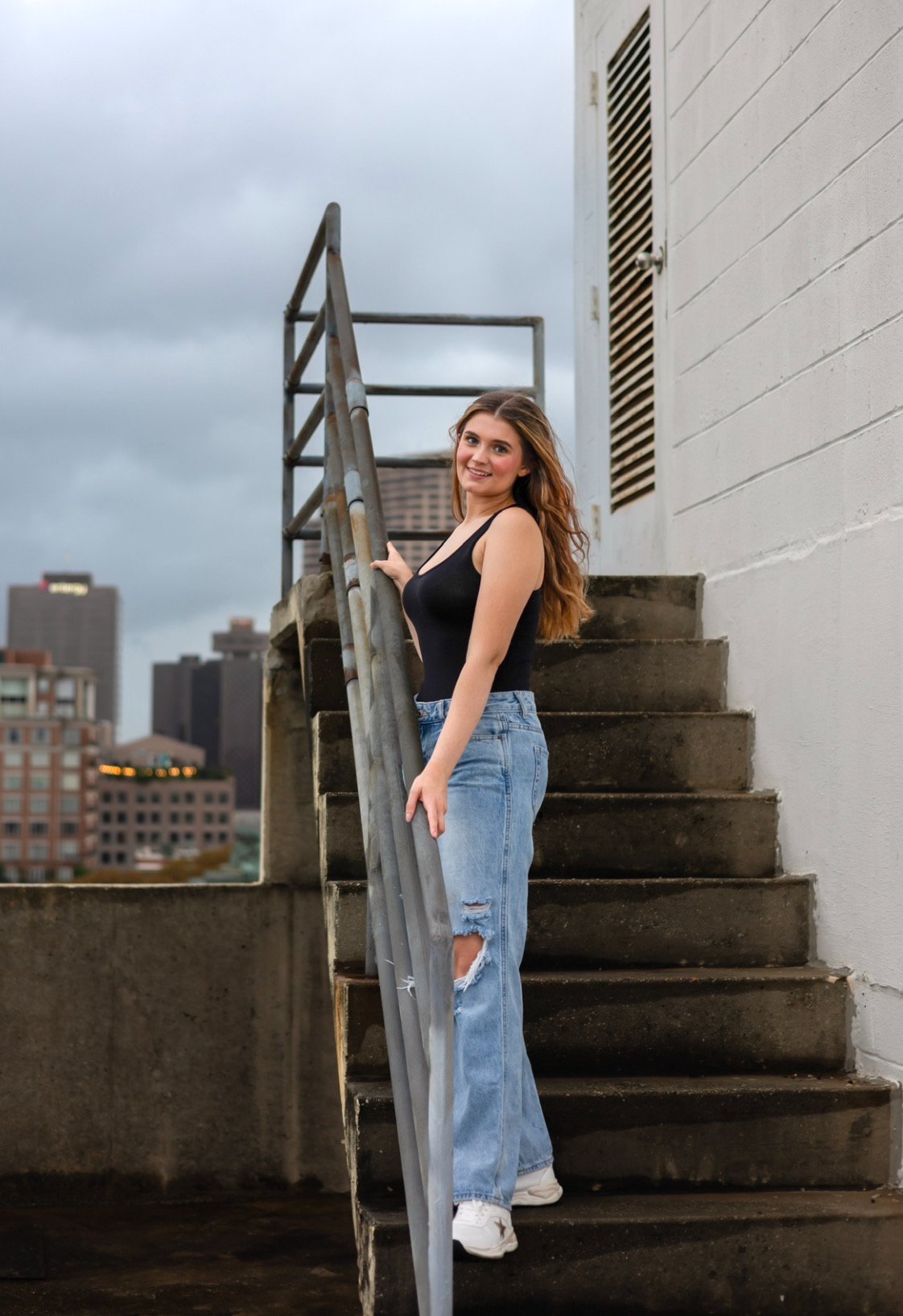 Woman in black top and ripped jeans standing on outdoor stairs on a rooftop with city skyline in background.