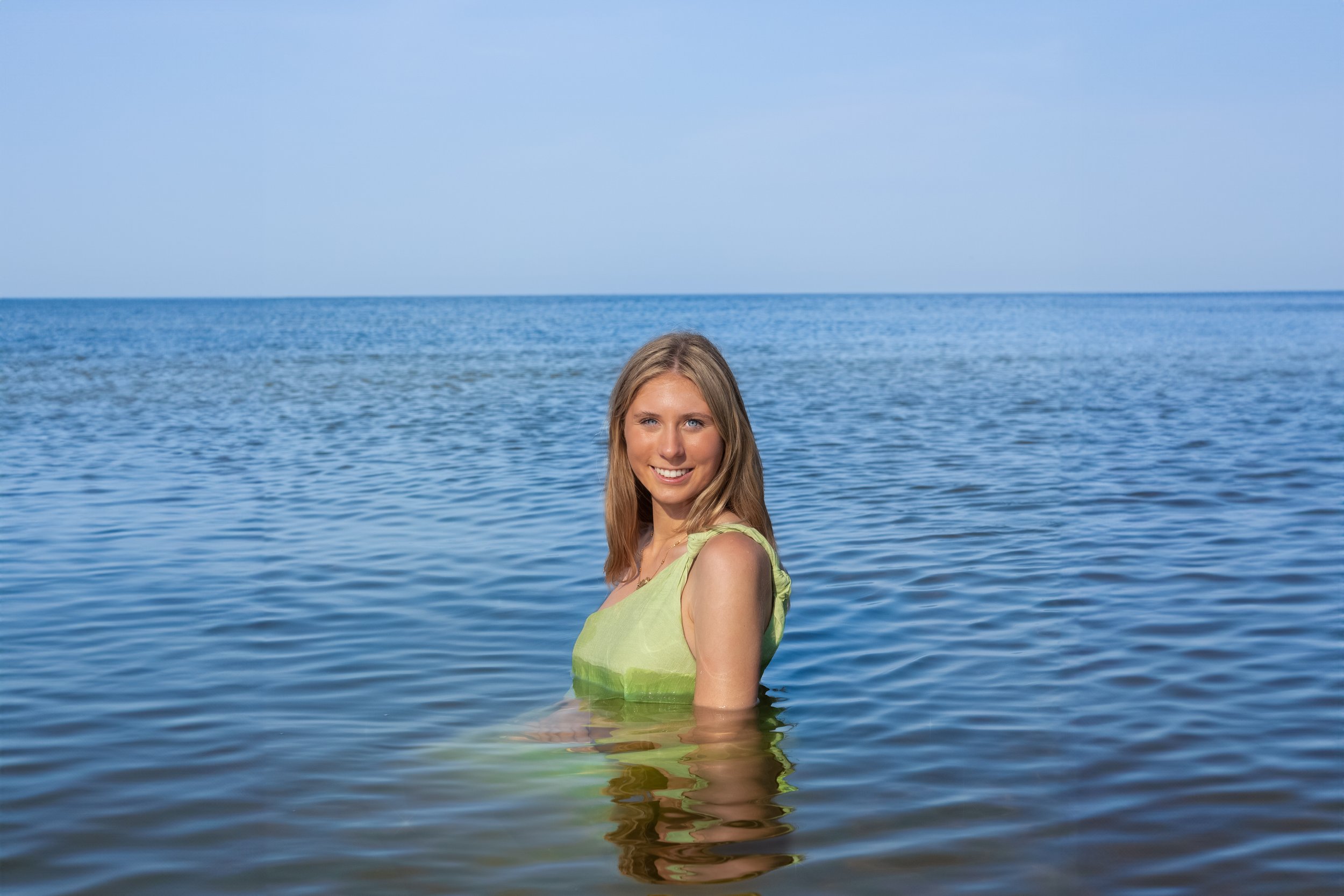 Woman smiling in a green dress standing in calm ocean water under a clear blue sky.