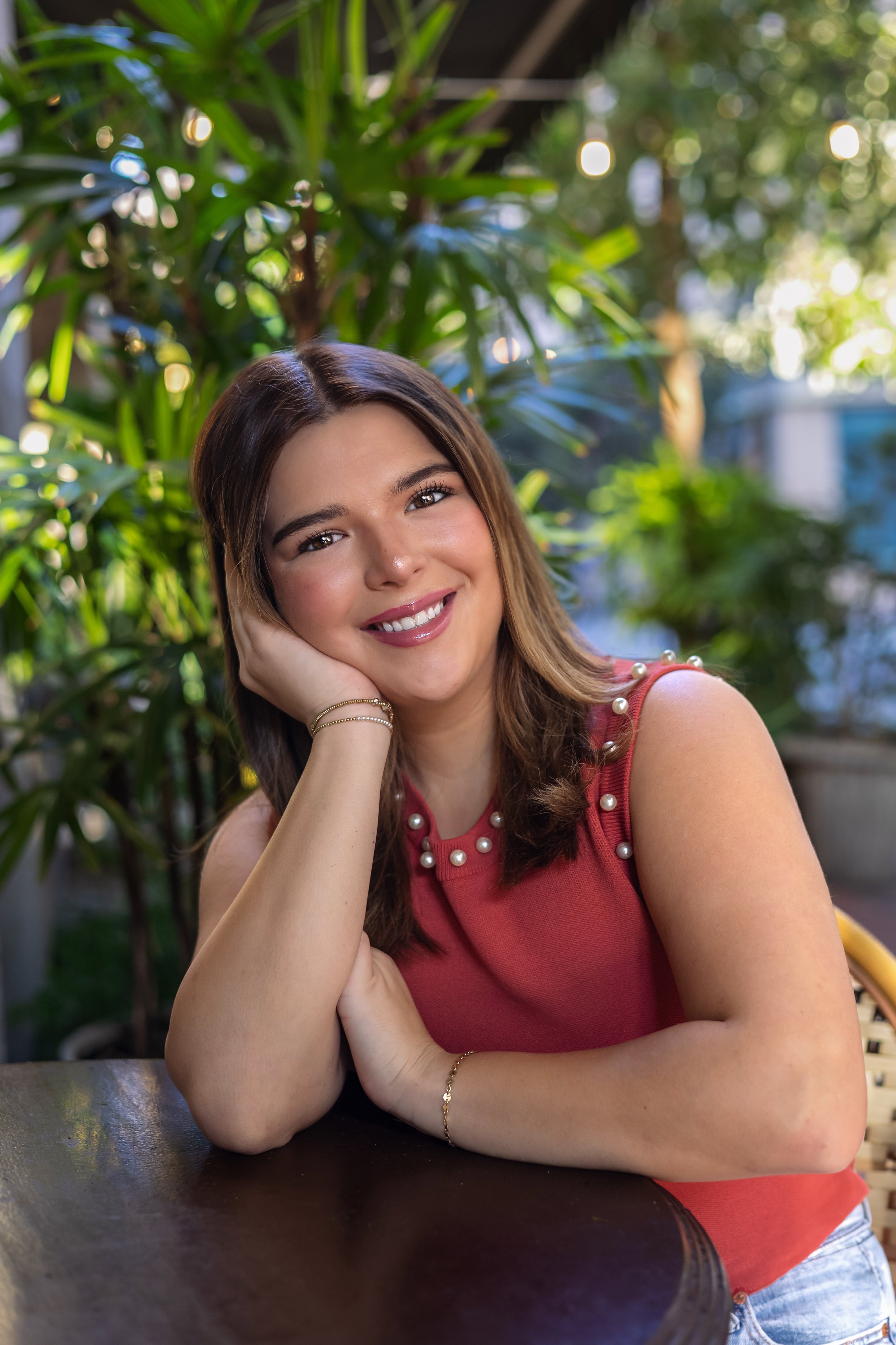 Smiling person seated at a table outdoors with plants in the background.