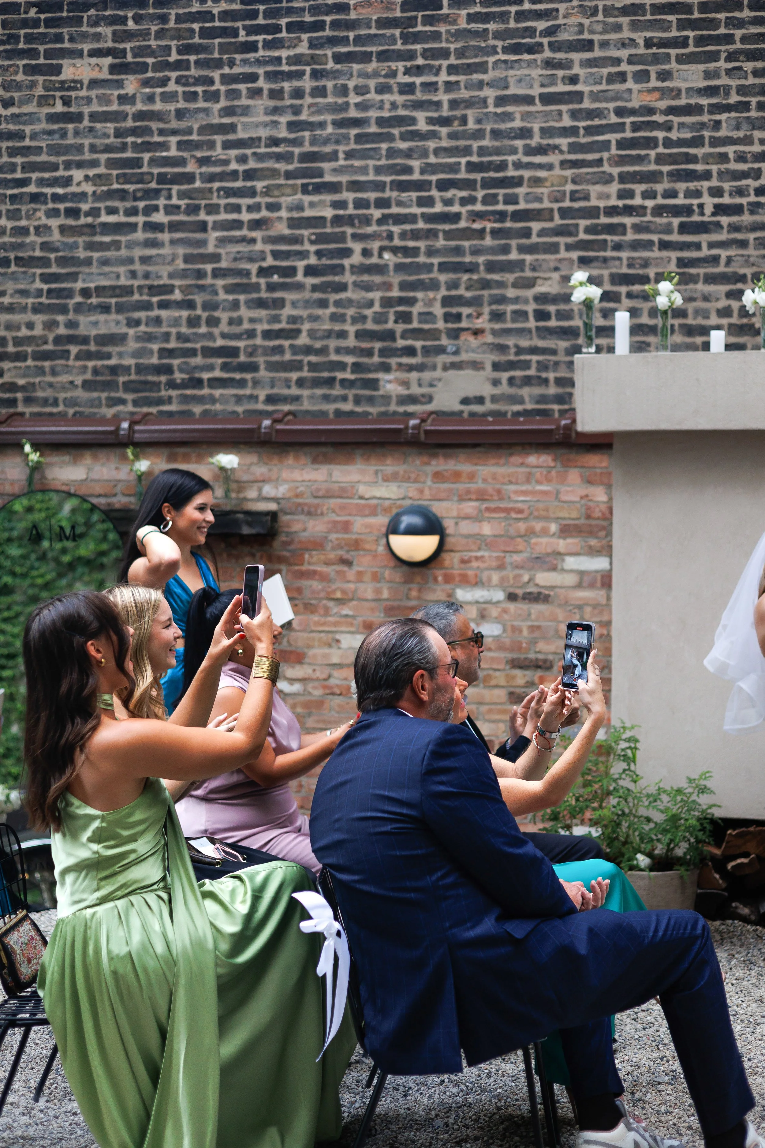 People sitting and standing outdoors, some taking photos, in front of brick and concrete wall with flowers.