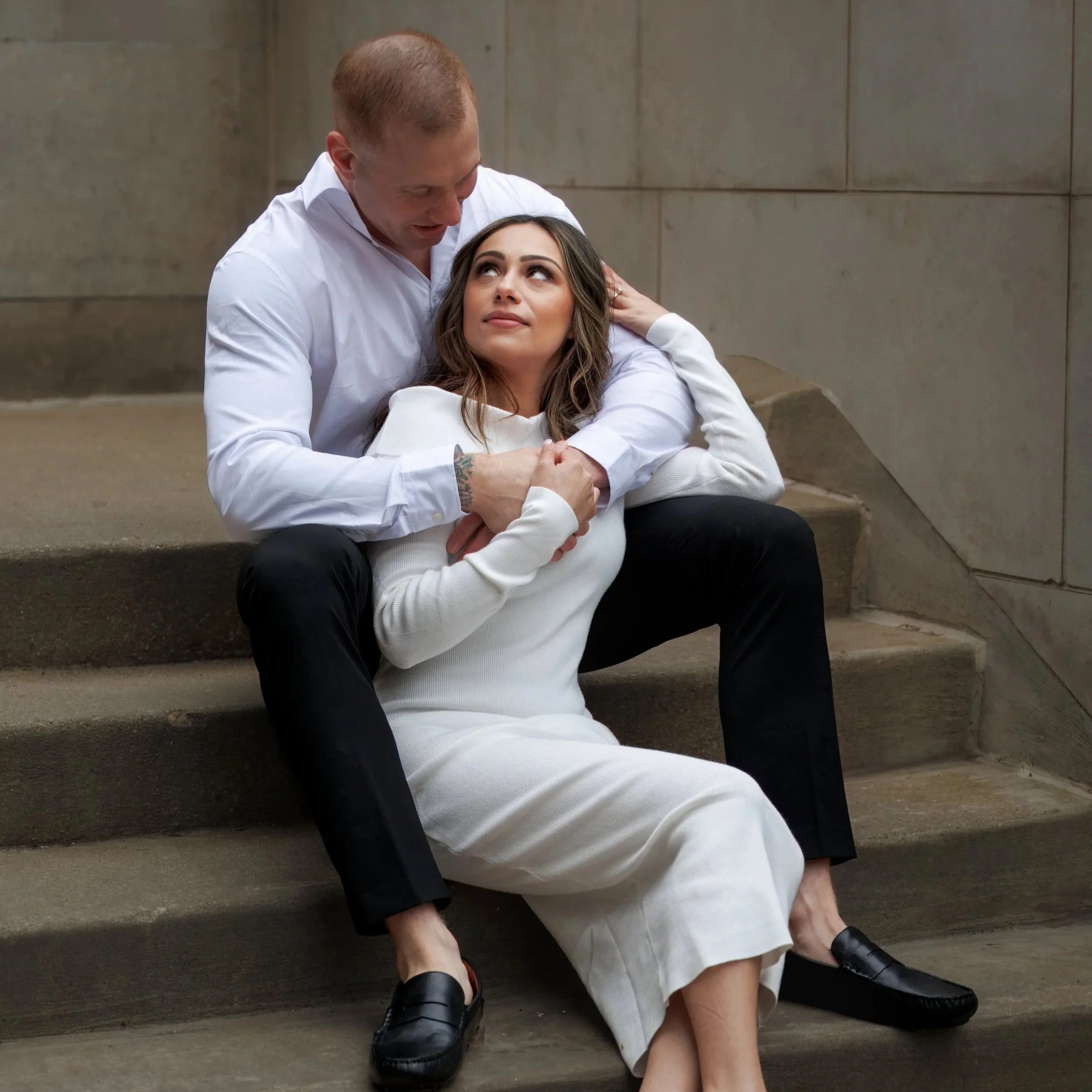 Romantic couples portrait on urban stone steps, man embracing woman in white dress during an intimate engagement-style photoshoot.