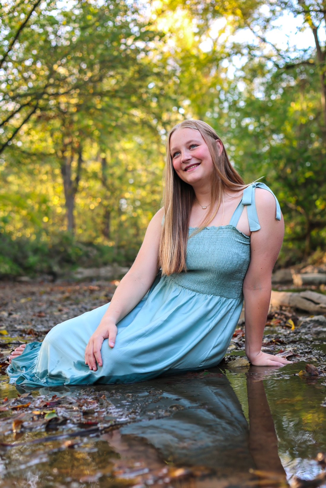 Woman in a light blue dress sitting by a creek in a forest, smiling, with sunlight filtering through the trees.