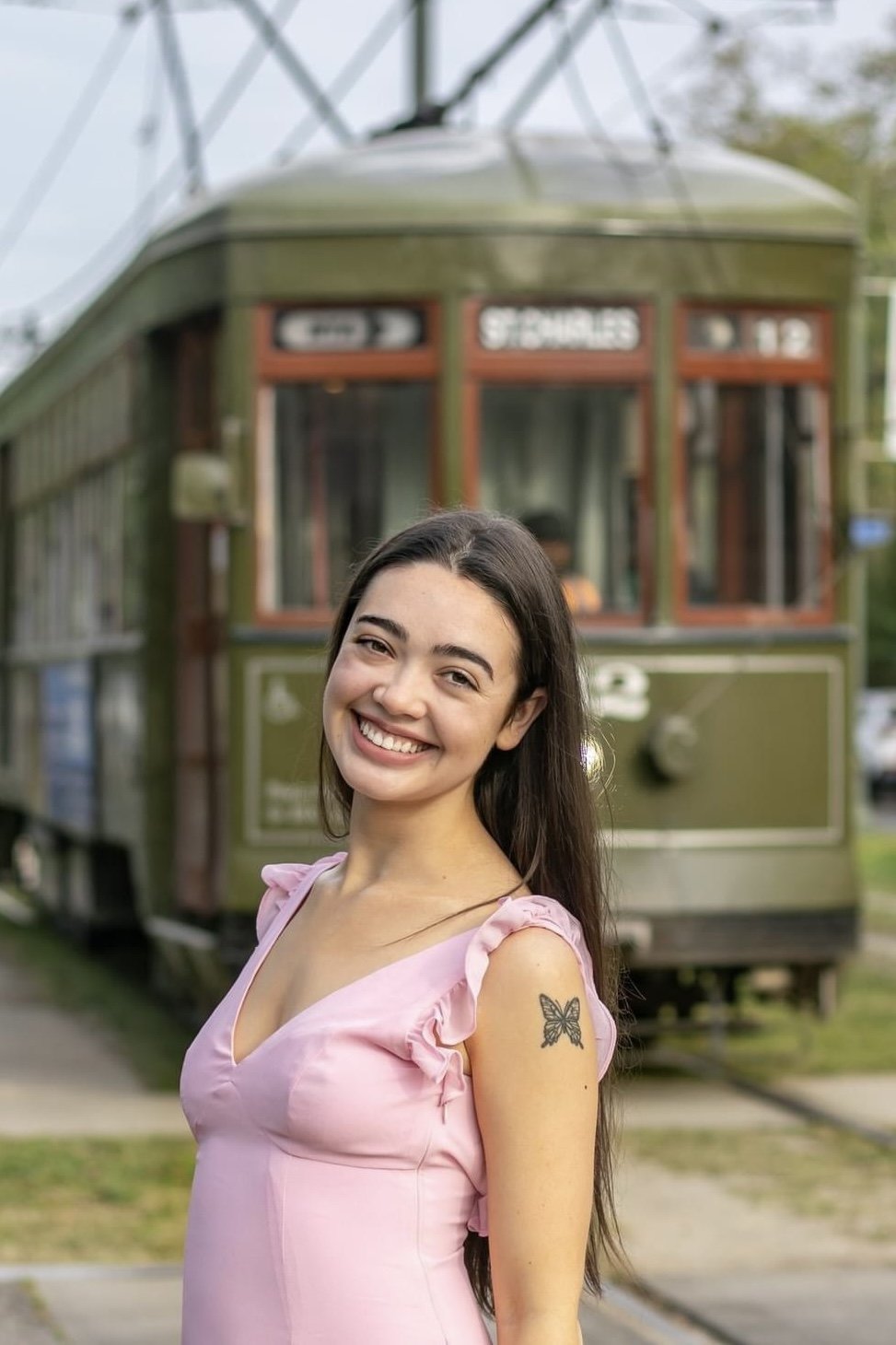 Smiling woman with long hair wearing a pink dress, standing in front of a historic green streetcar with a butterfly tattoo on her shoulder.