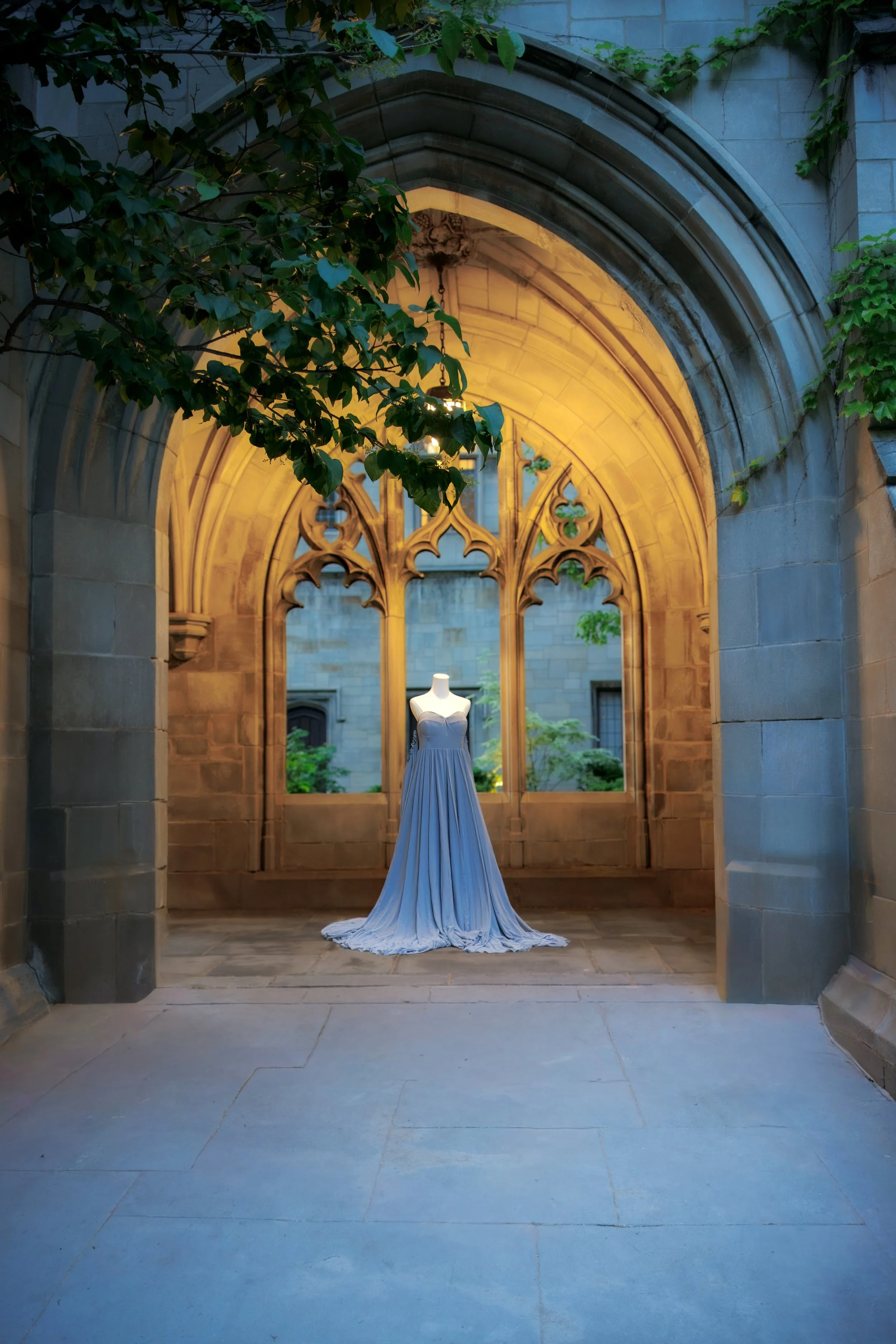 A white wedding dress displayed on a mannequin inside a historic stone archway illuminated with warm lighting, greenery hanging around the arch.