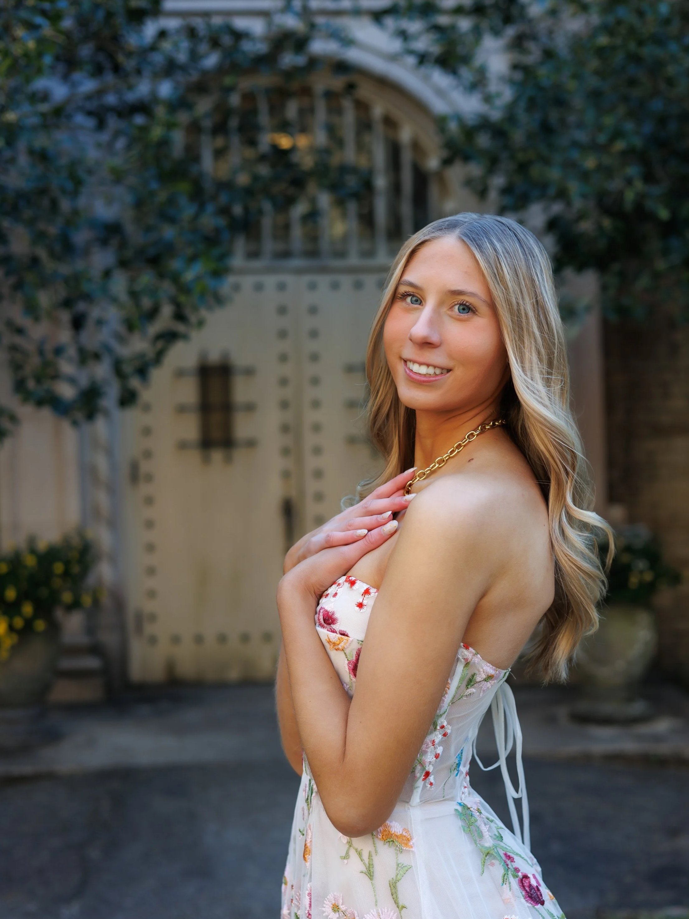 Chicago wedding photographer captures a young woman with long blonde hair and blue eyes smiling outdoors in front of a decorative door and greenery, wearing a strapless floral dress and a gold chain necklace.
