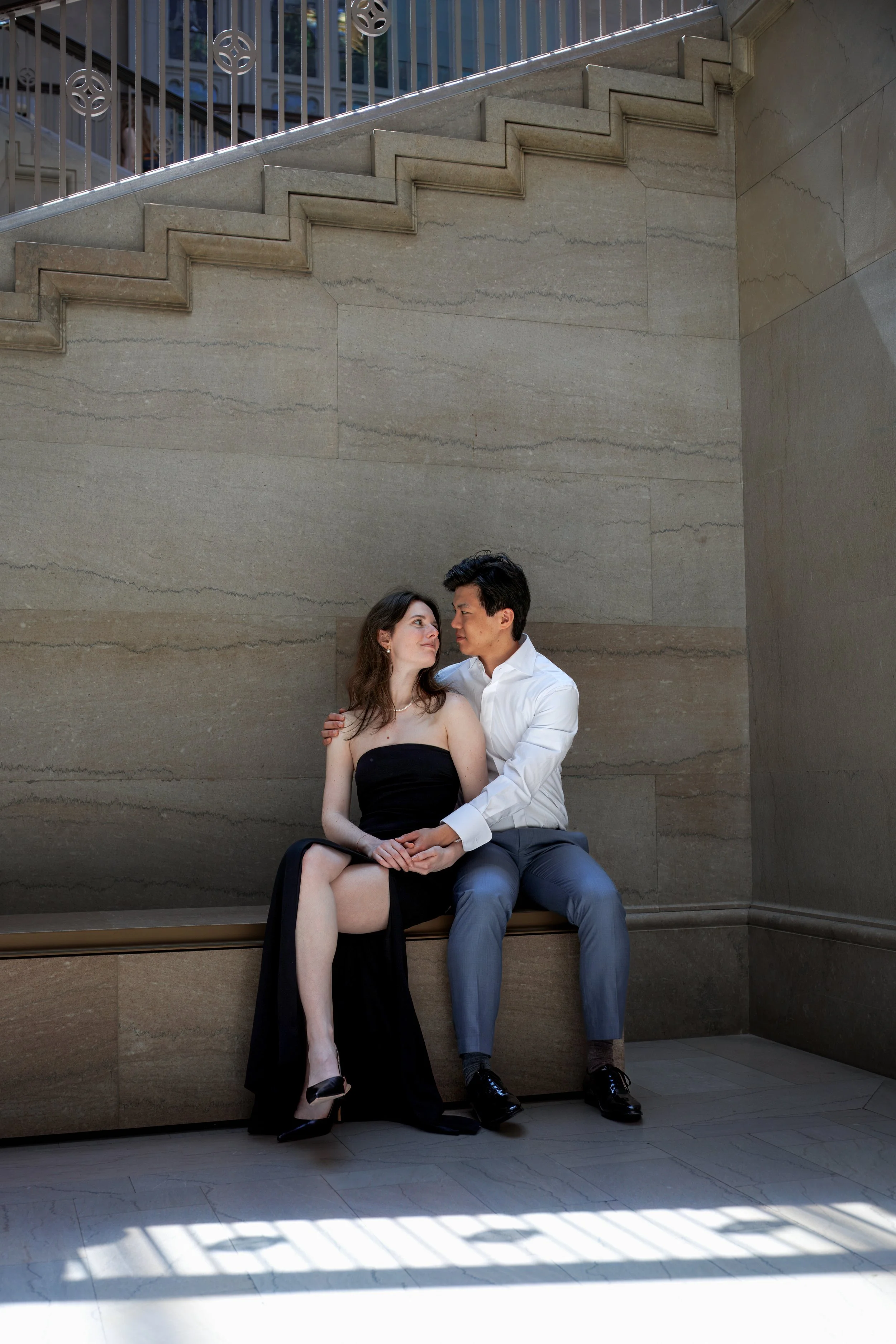 Couple seated beneath the staircase inside the Art Institute of Chicago, sharing a quiet moment in soft natural light against marble walls.