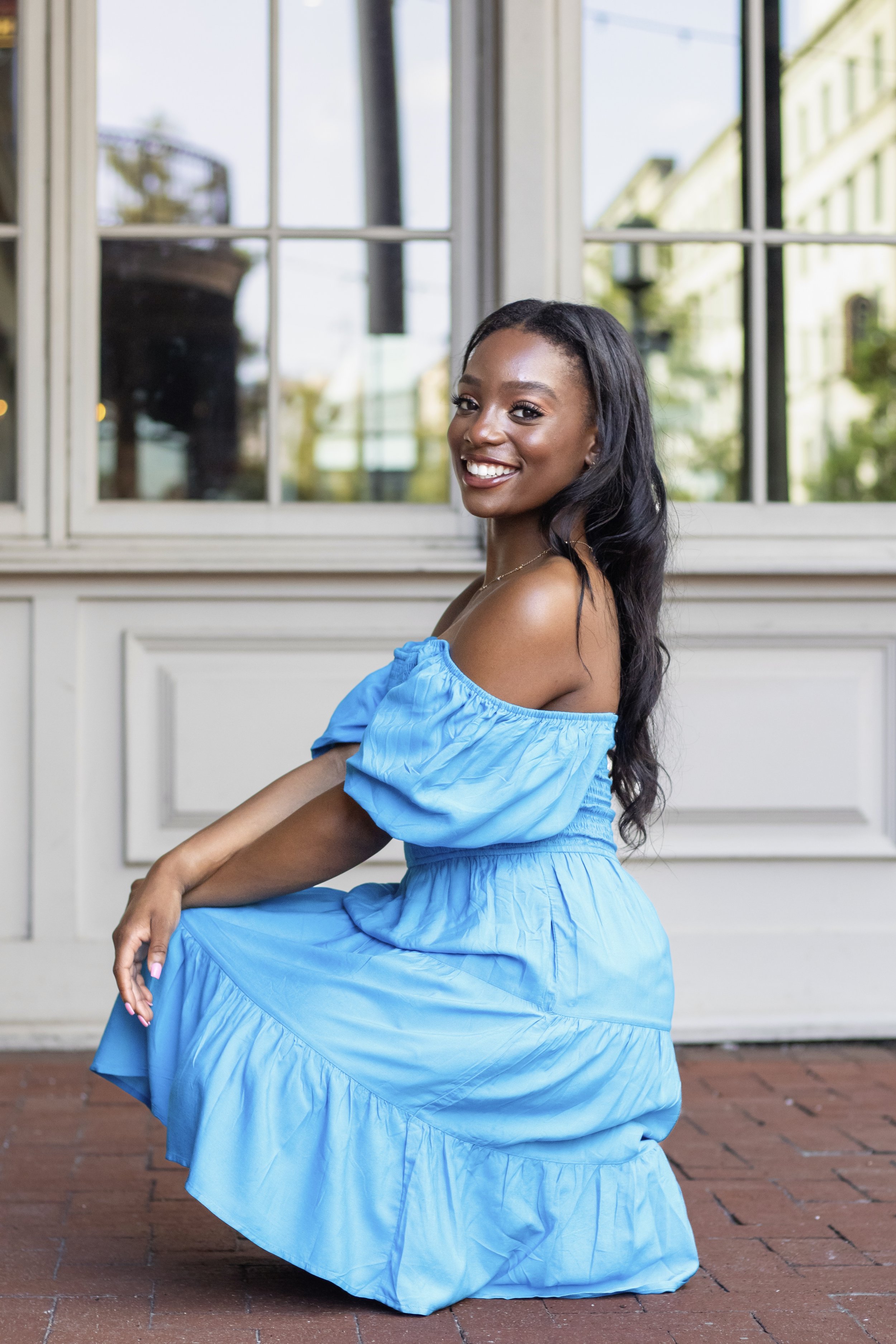 Woman in blue dress kneeling and smiling outdoors, in front of windows with reflections.