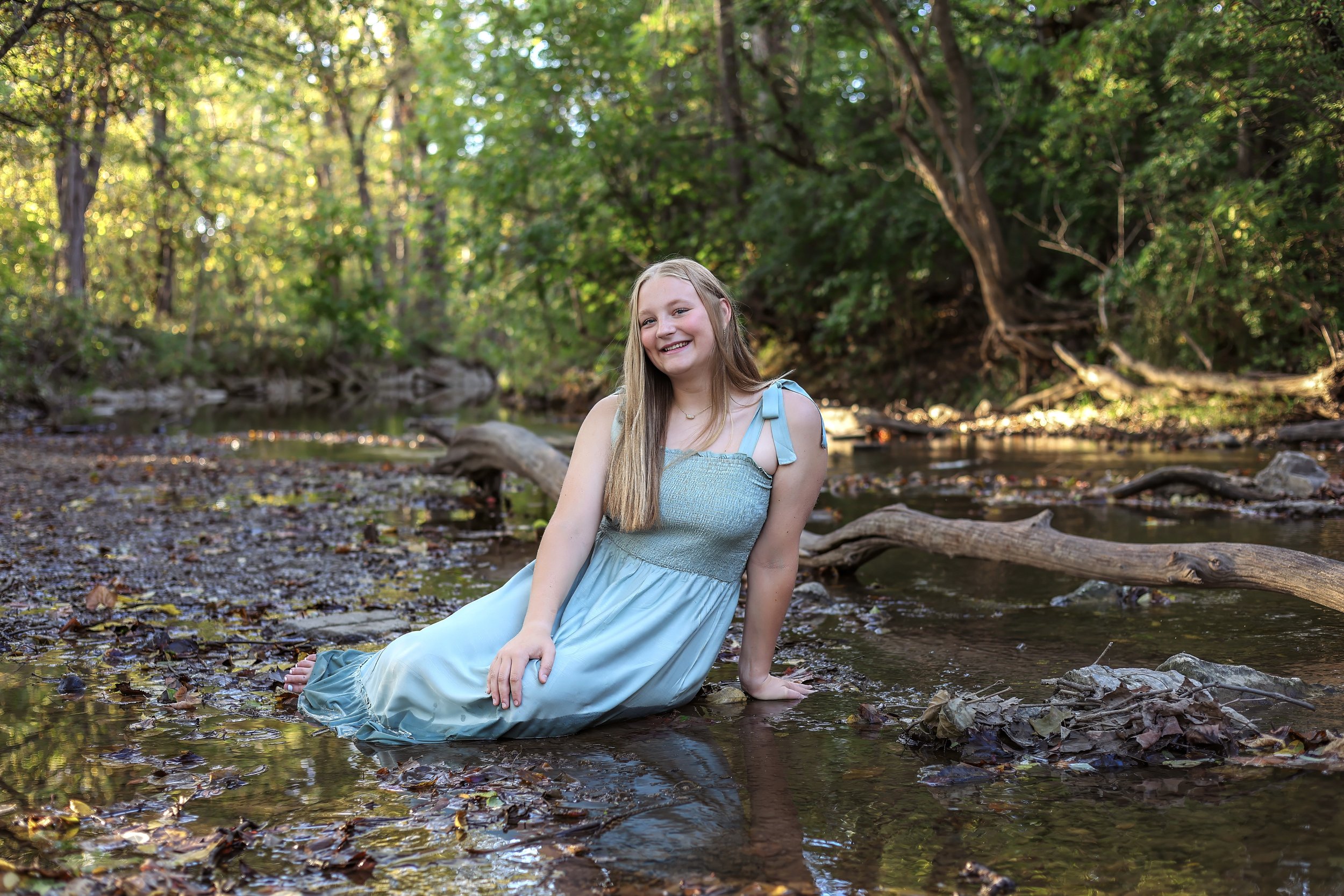 Smiling person in a light blue dress sitting in a forest stream, surrounded by trees and fallen leaves.