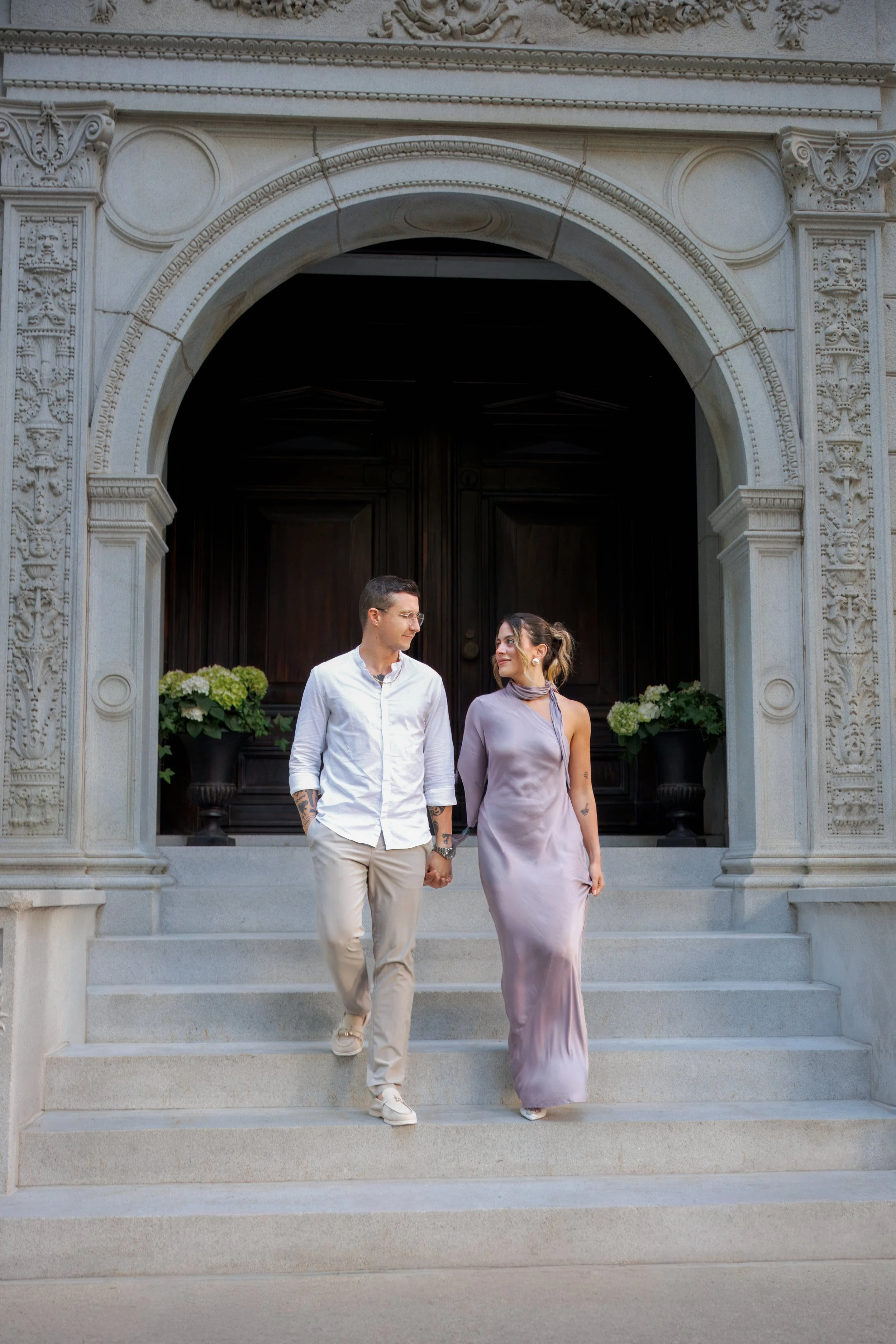Engaged couple holding hands and walking down the steps of an ornate stone building while looking at each other, photographed during a romantic engagement session