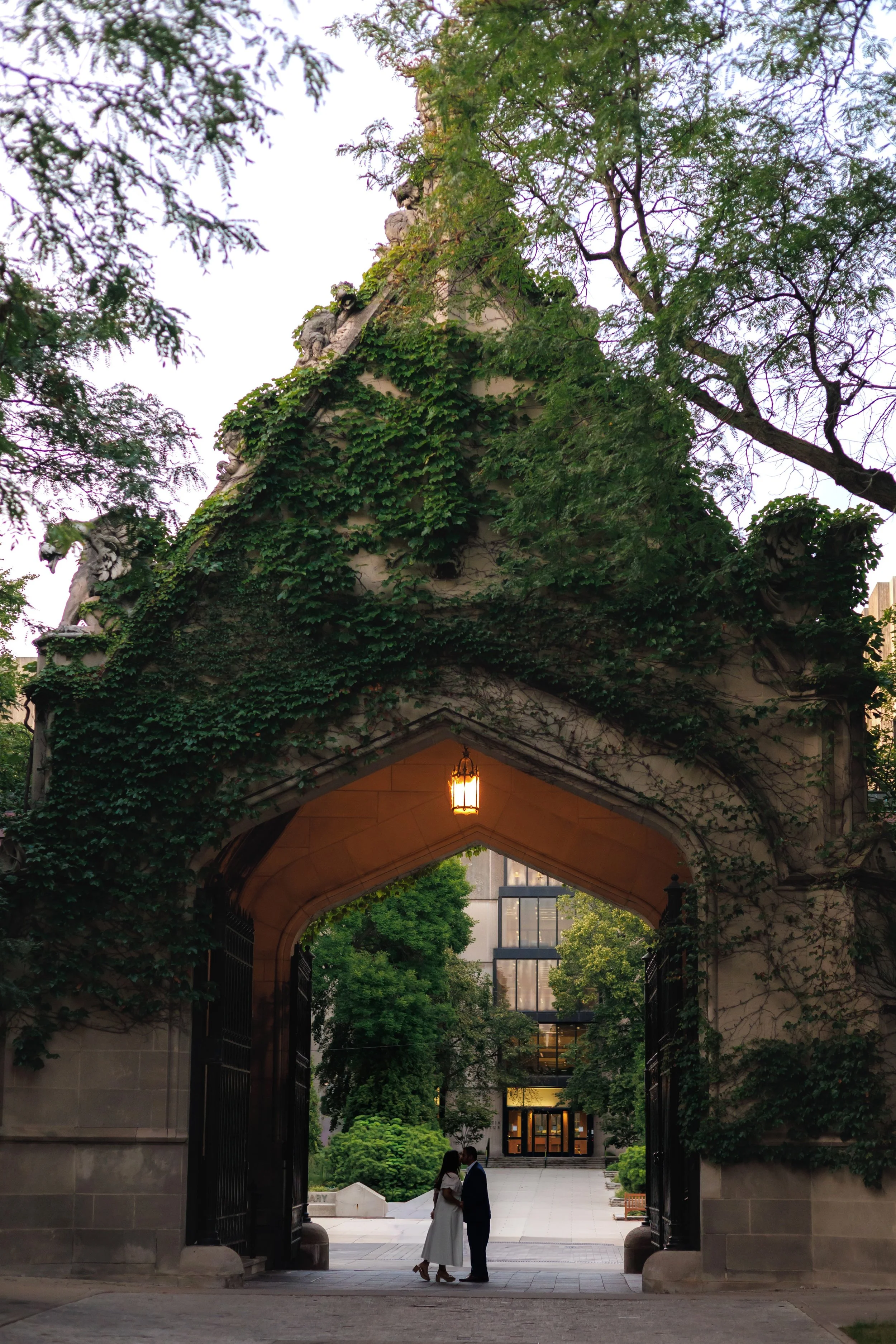 Chicago wedding photographer captures a bride and groom standing close together in front of a large open gateway with ivy-covered stone walls and trees, a modern building with glass windows in the background, and a hanging lantern