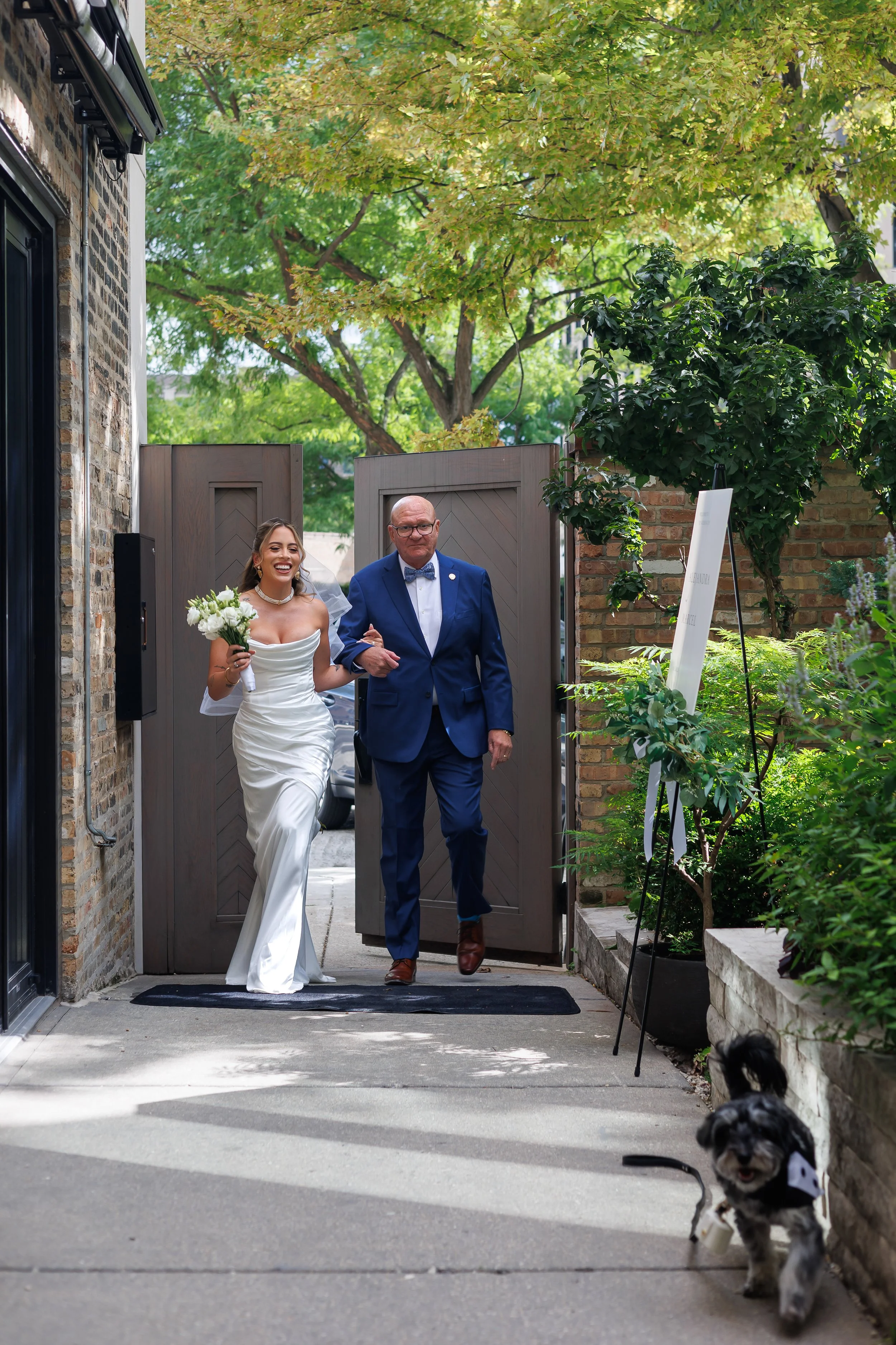 A bride in a white wedding dress walking out of a gate alongside an older man in a blue suit. The bride is holding a bouquet of white flowers and smiling.