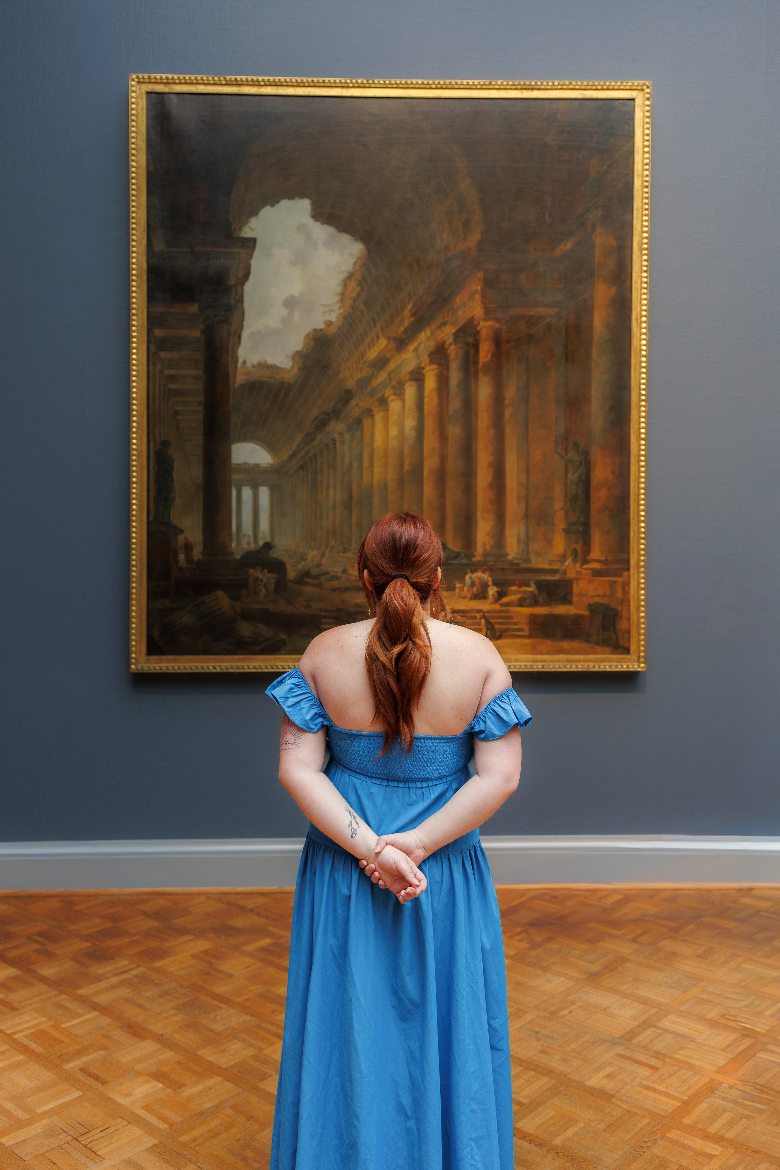 A woman in a blue dress stands with her back to the camera, observing a large classical painting in the Art Institute of Chicago. The painting depicts an ancient architectural scene with columns, statues, and clouds. 
