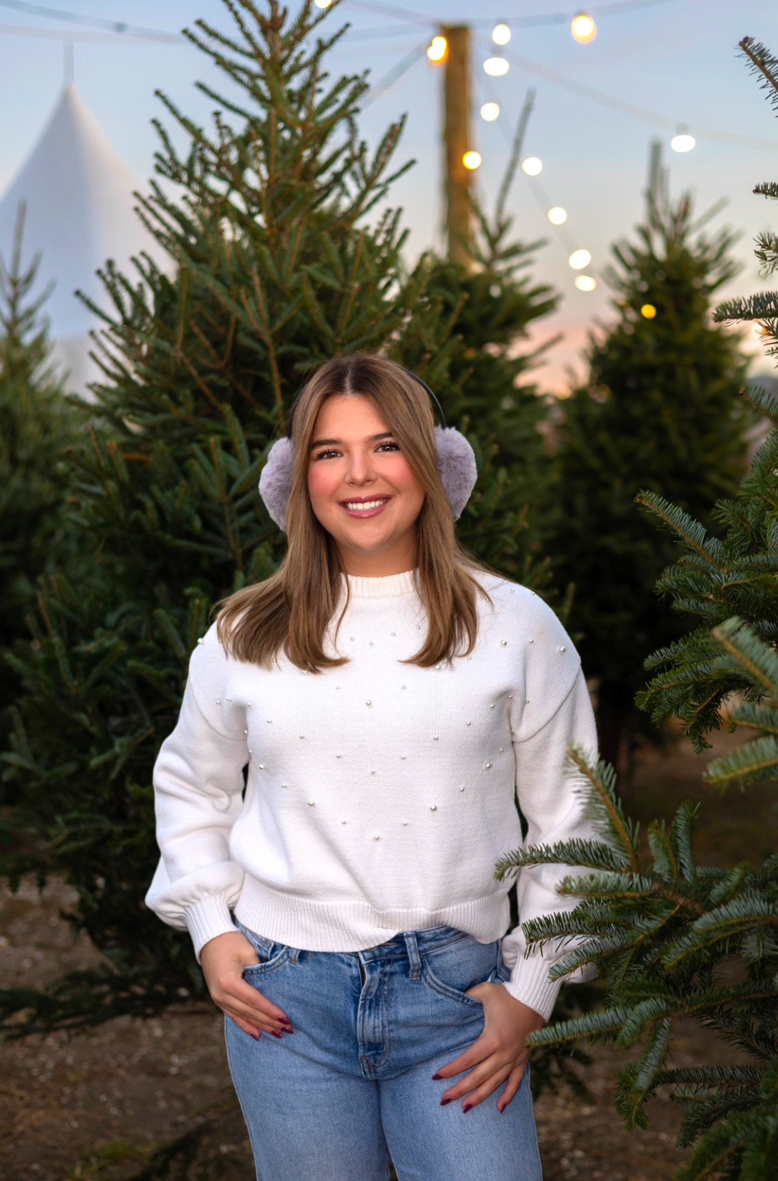 A person wearing a white sweater and earmuffs stands among Christmas trees with decorative lights in the background.