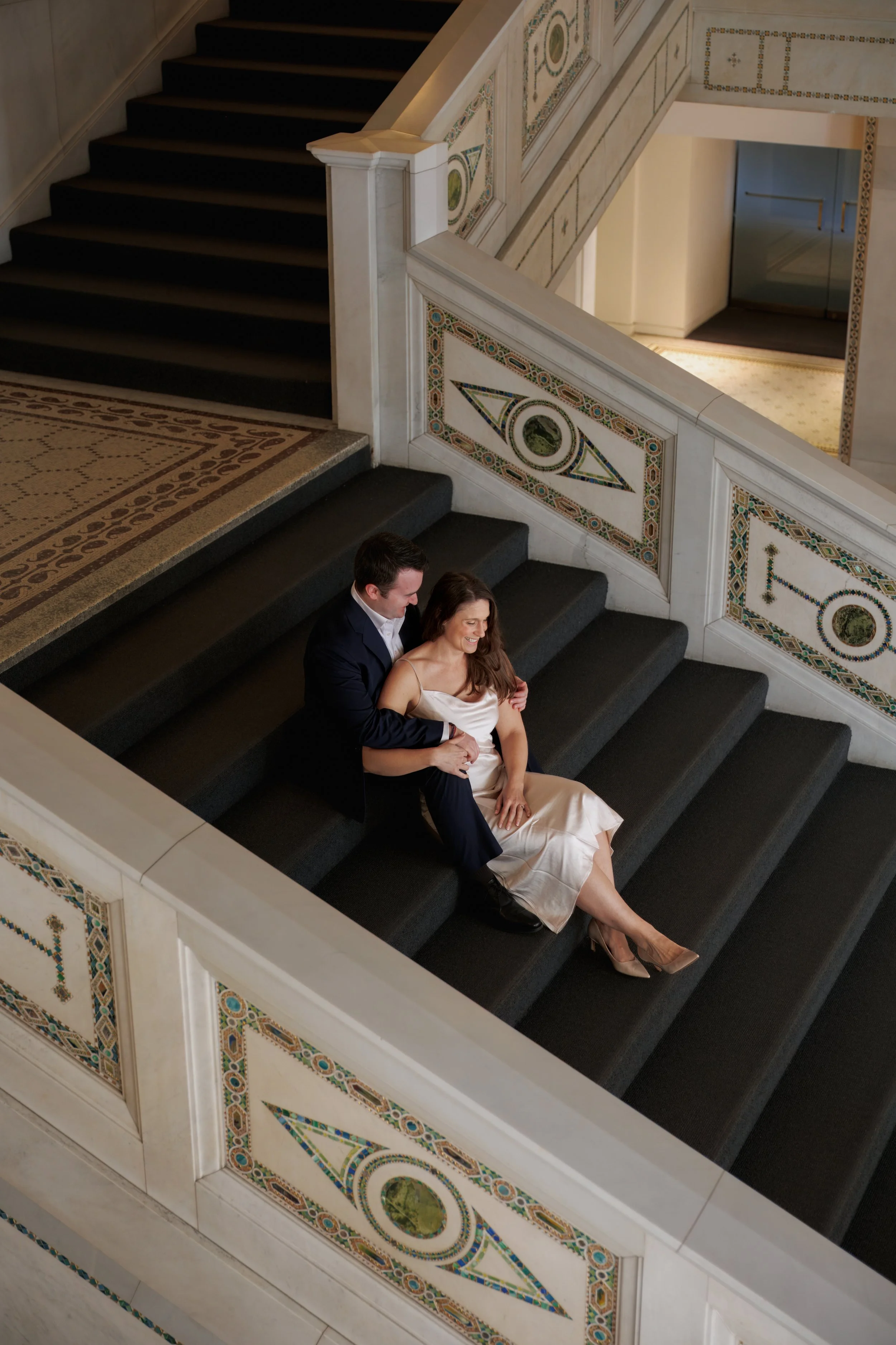 Couple sitting close together indoors and interacting naturally during an intimate engagement session with soft, neutral tones