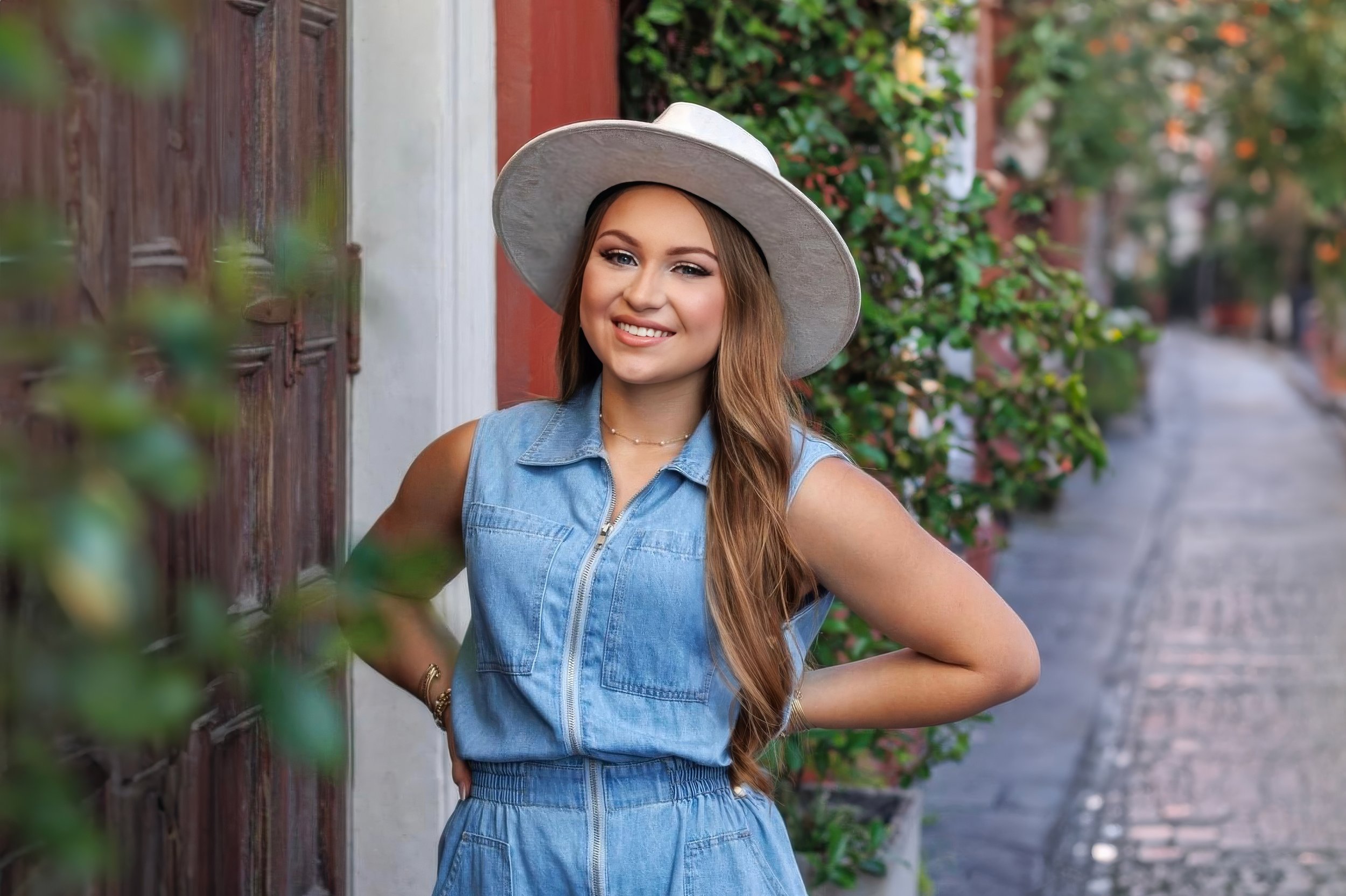 Woman in denim outfit and white hat standing on a cobblestone street with green foliage in the background.