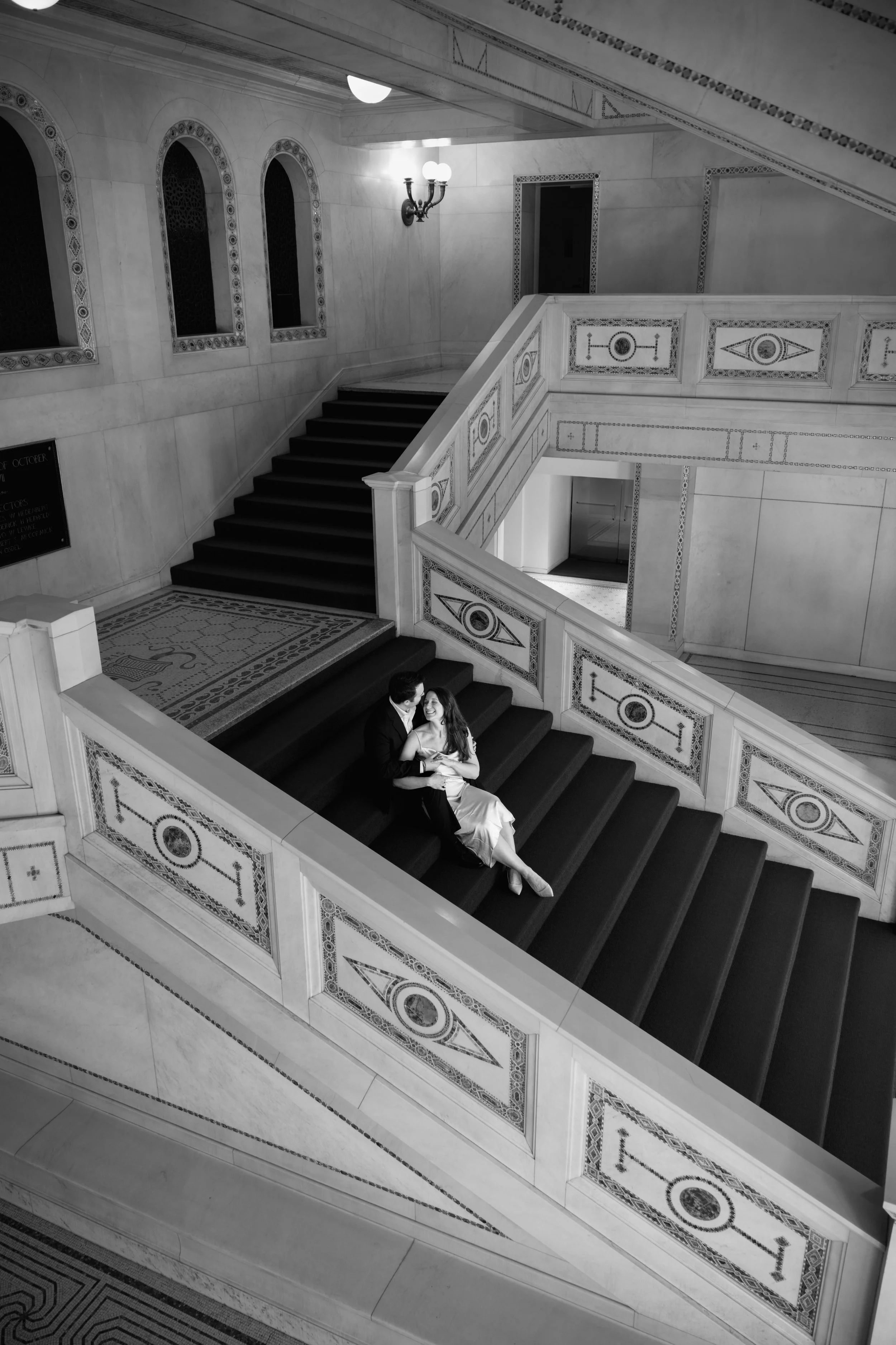 A man and woman sitting together on a staircase in an elegant, historic building with ornate wall and railings, in black and white.
Chicago wedding photographer