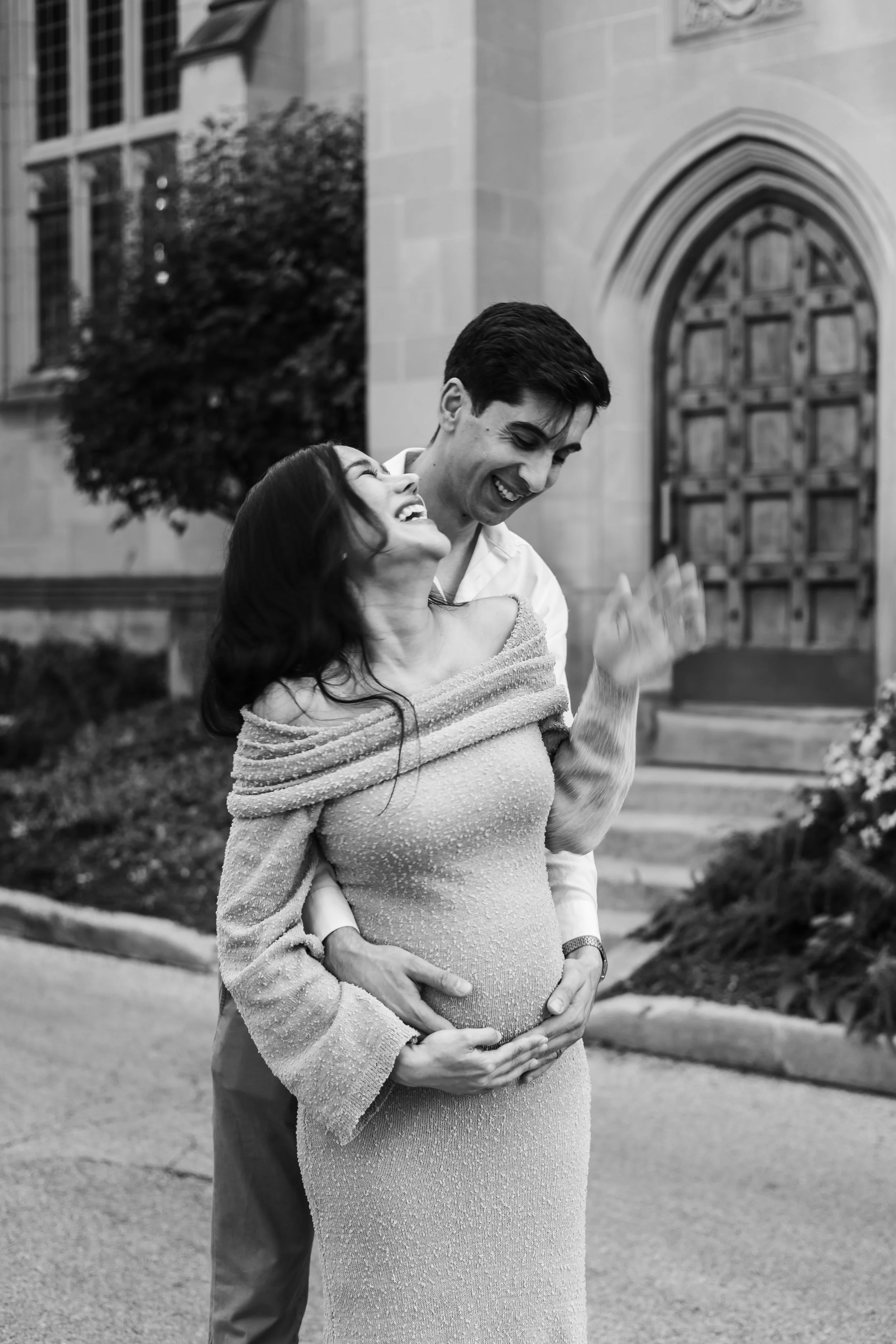 A joyful pregnant woman and a man are holding each other in front of a stone building with an arched wooden door, sharing a moment of happiness. Chicago wedding photographer