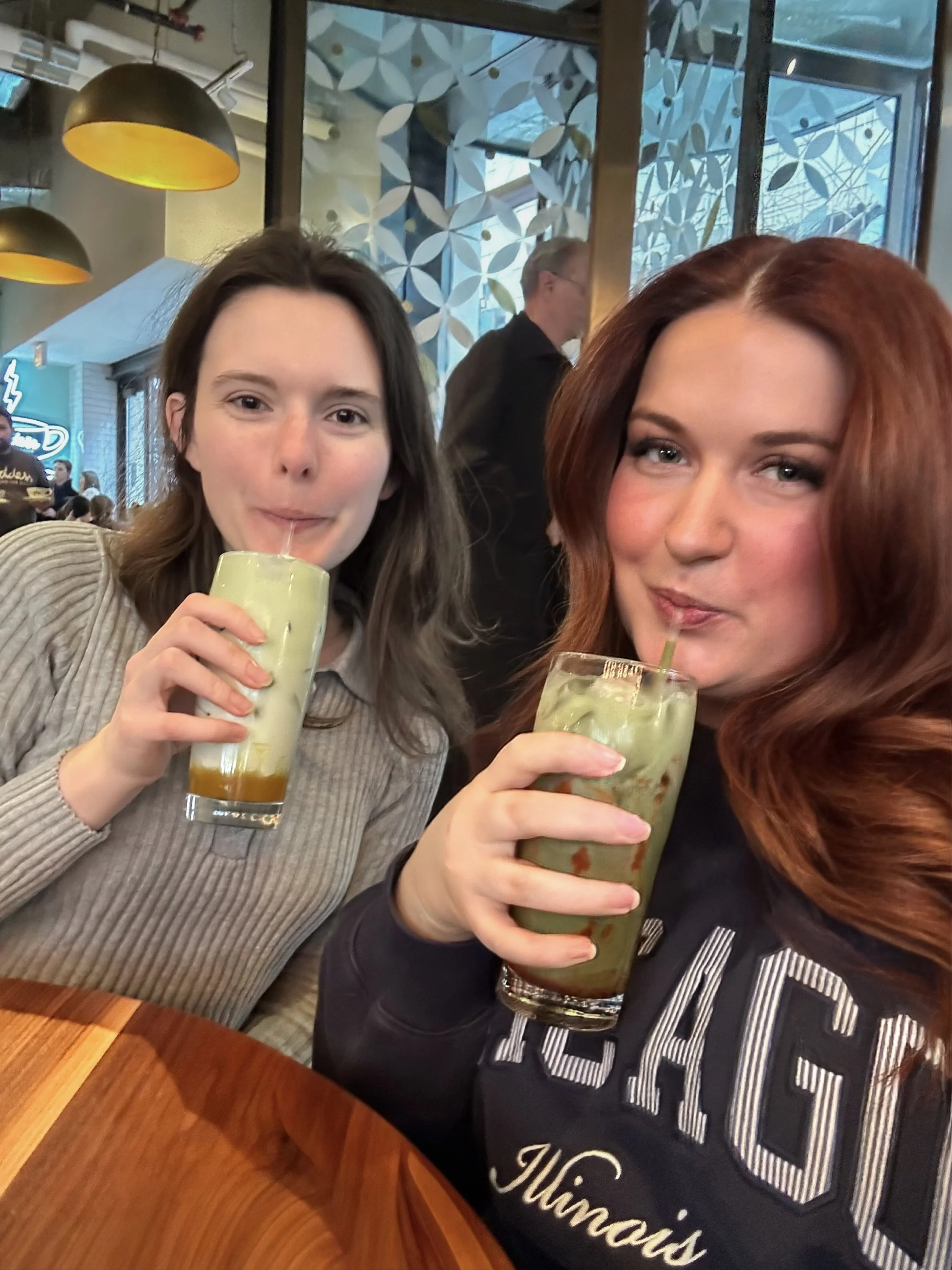 Two women sitting at a table in a restaurant or café, drinking iced green drinks with straws, with a window and decorative wall in the background. Chicago wedding photographer