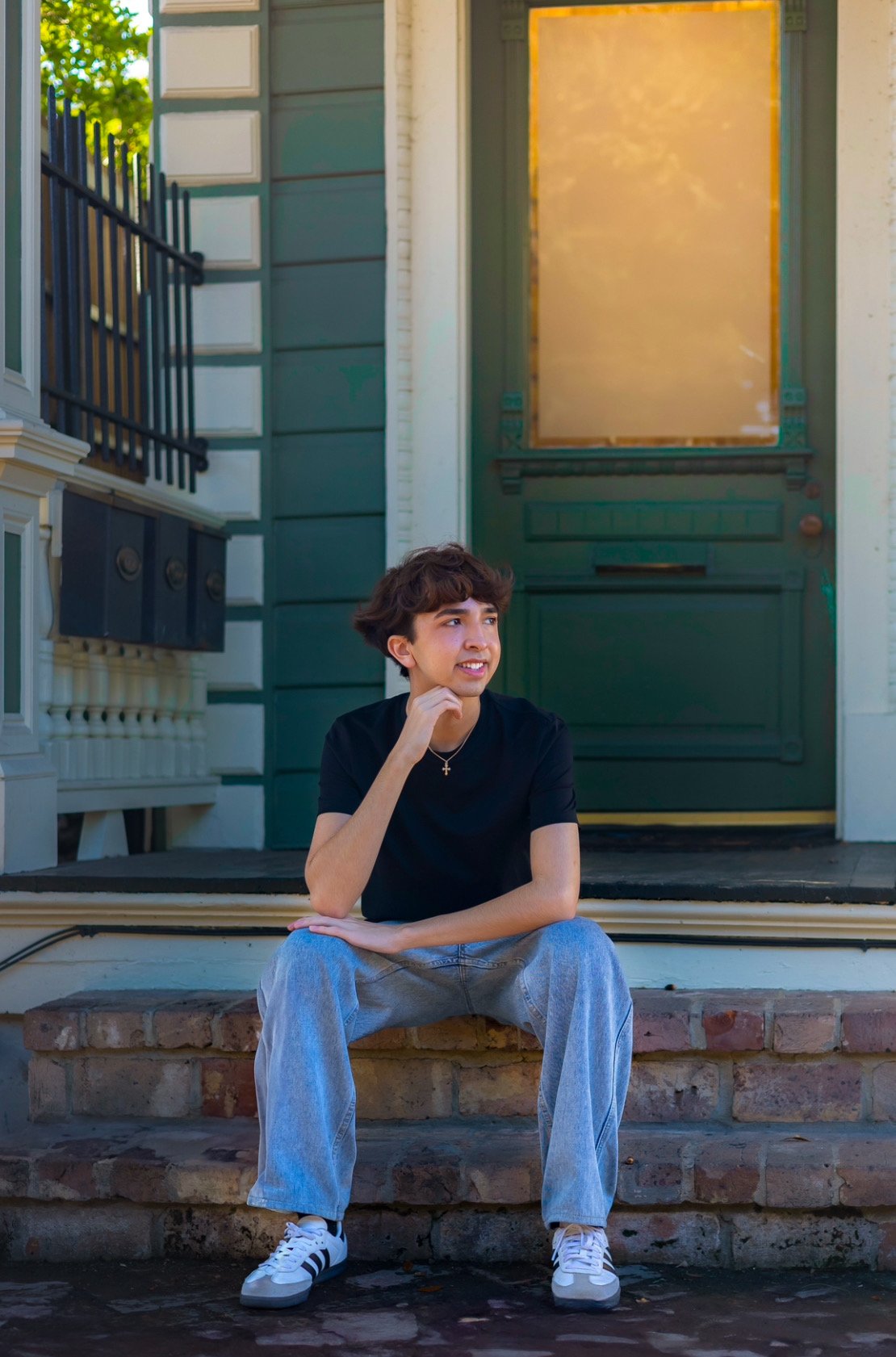 Person sitting on steps in front of a door, wearing a black shirt and jeans, with hand on chin.