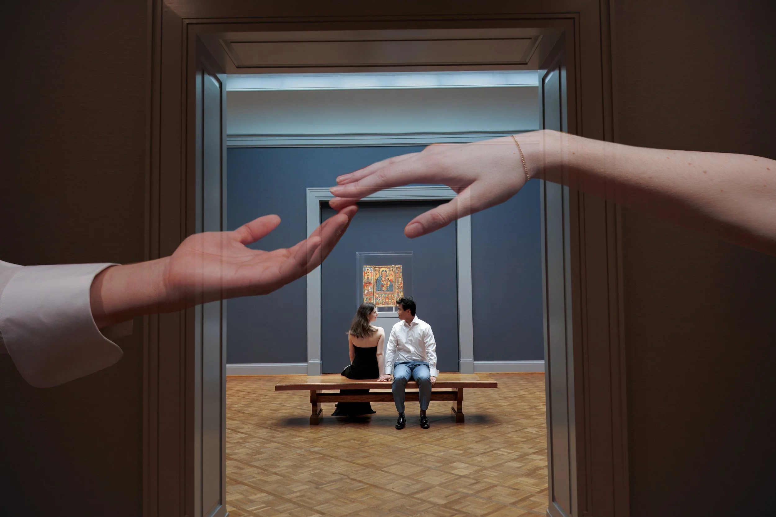 A still from a film or TV show of a man and a woman seated on a bench, facing each other, in an art gallery. The scene is viewed through a doorway with a framed opening. Their hands are touching, and there is a religious icon on the wall behind them.