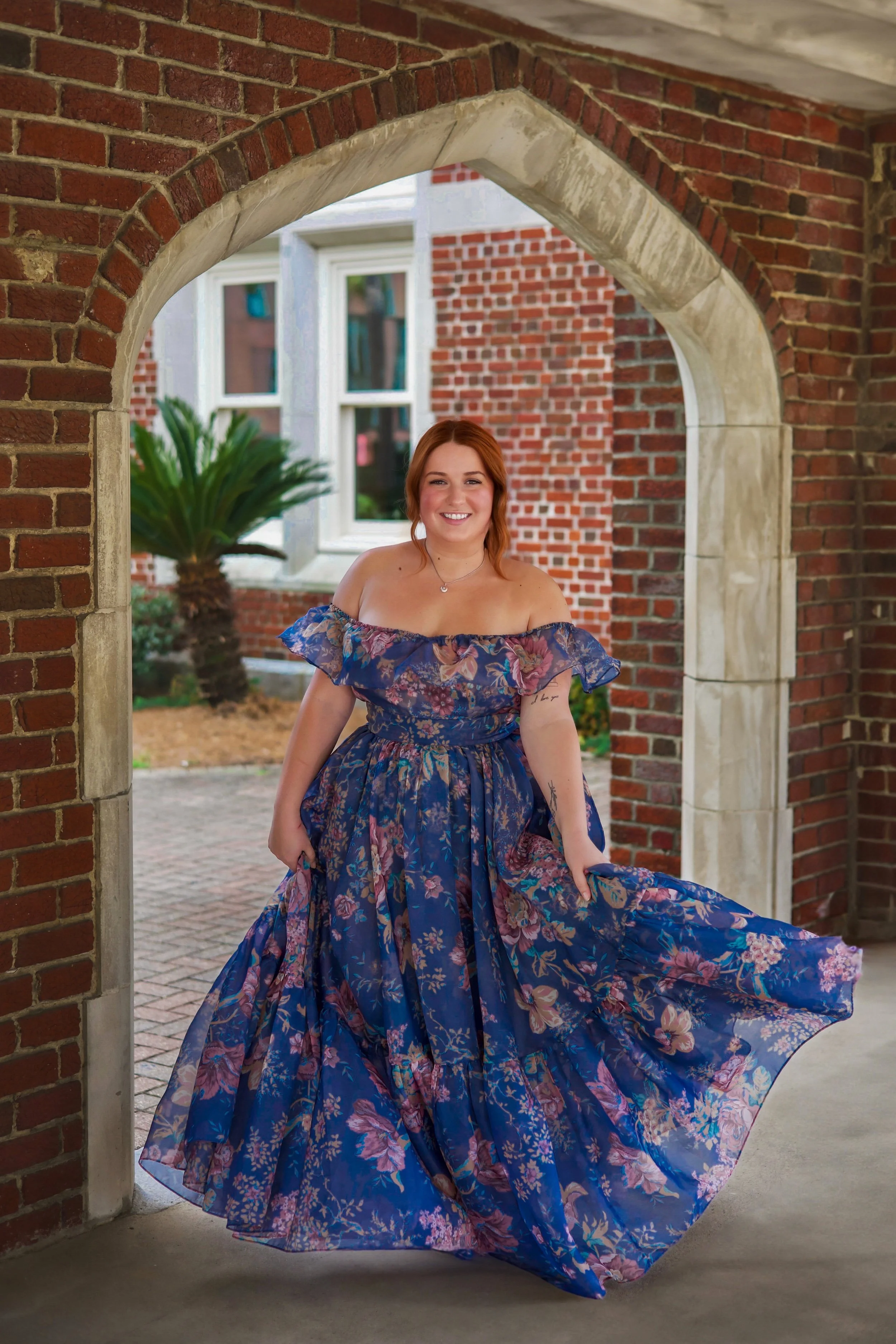 A photographer in a long, blue floral dress standing in an arched brick entryway in Chicago, smiling.