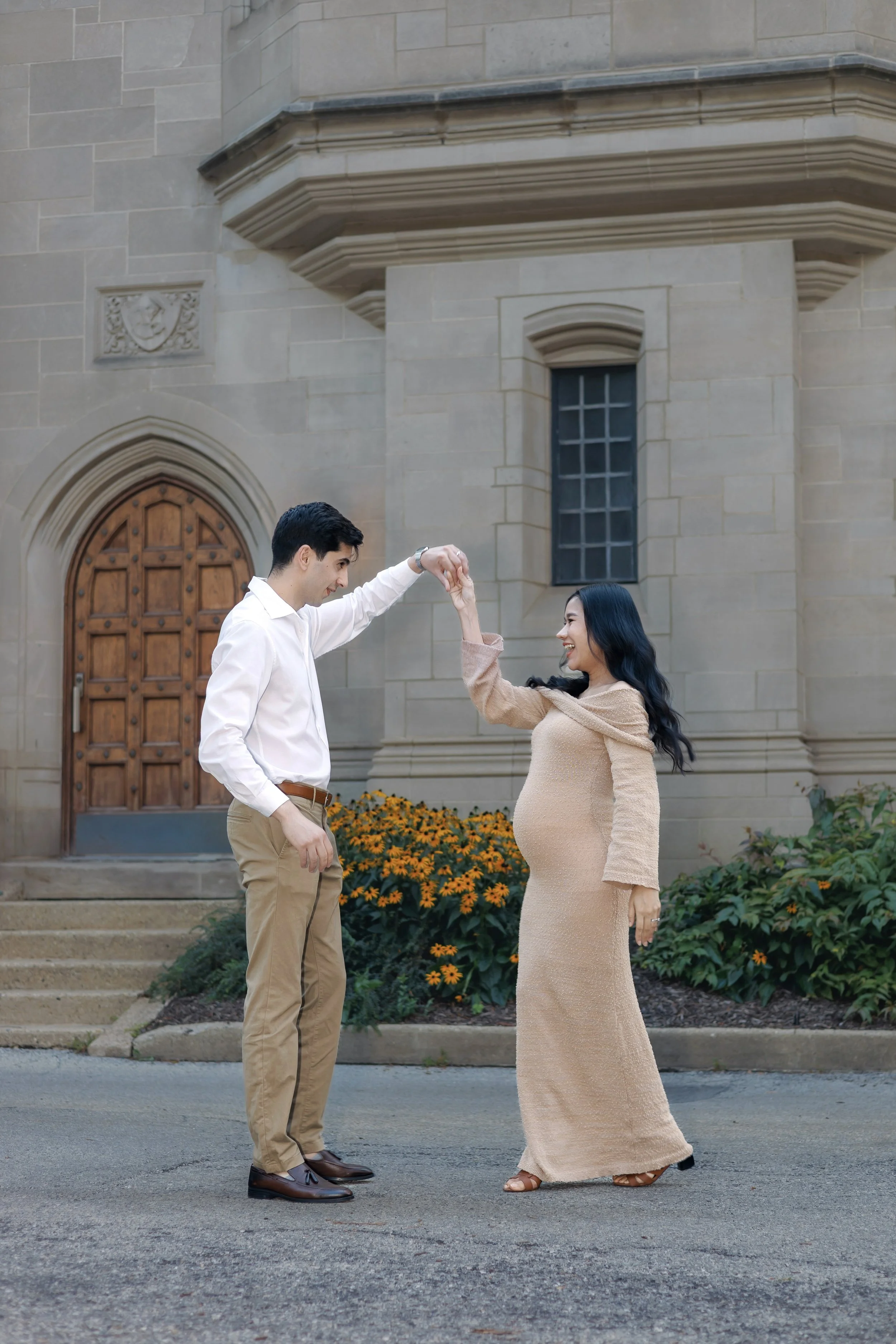 Chicago maternity photographer capturing a couple dancing hand in hand in front of a castle like building, soft light creating a warm and romantic maternity moment