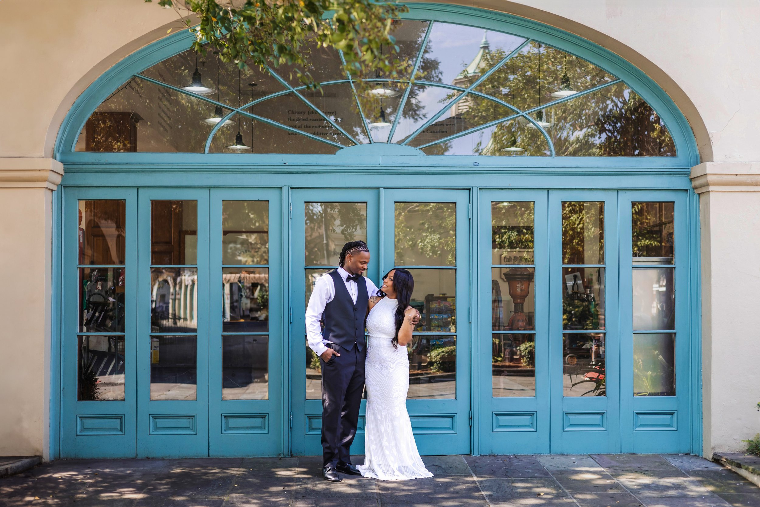 A couple is sitting in a silver canoe on a calm river, surrounded by lush greenery and a stone bridge in the background. The man is embracing the woman, who is relaxed with her legs on the edge of the canoe.