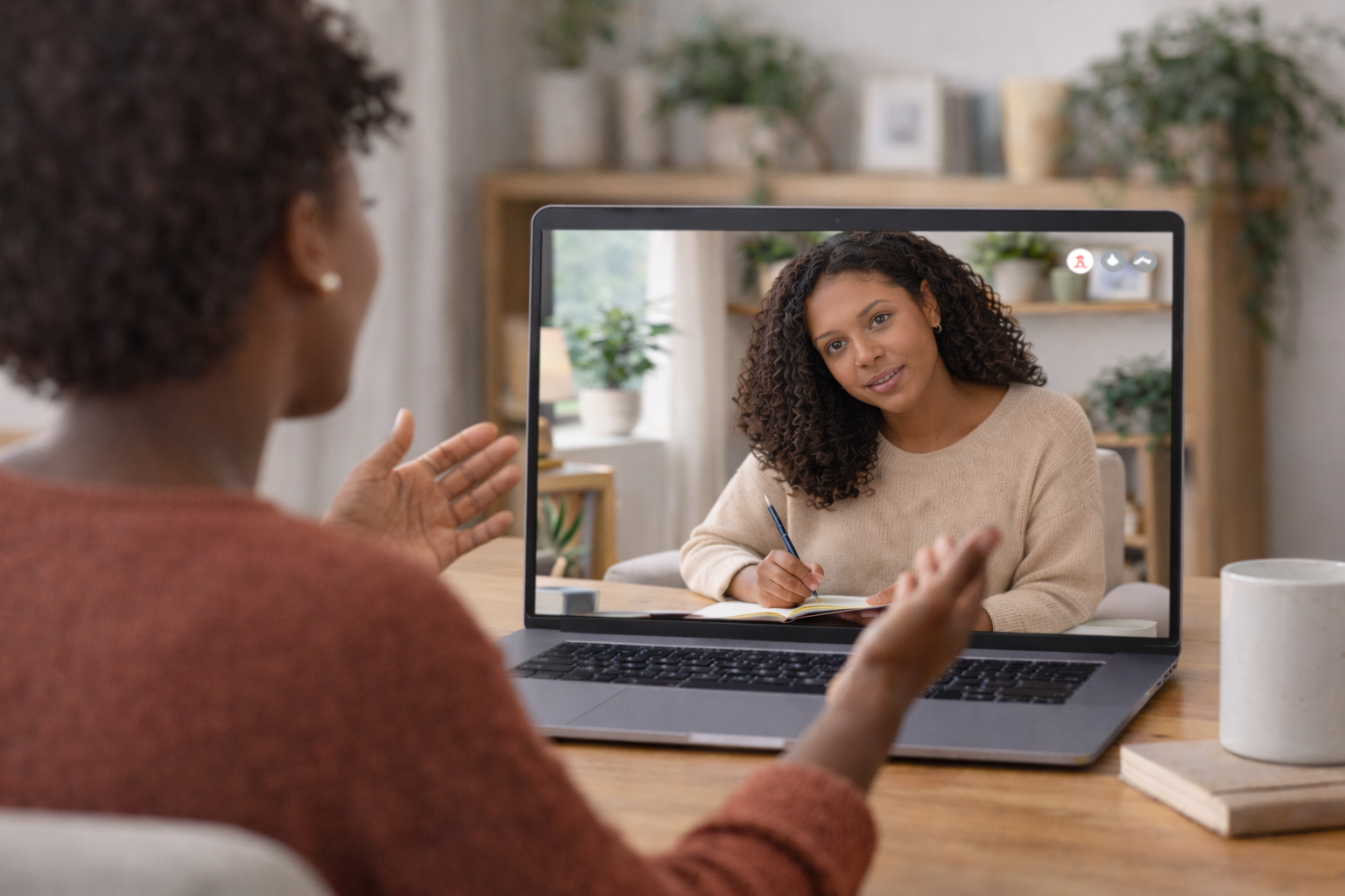 Black woman participating in a virtual therapy session, speaking with expressive hand gestures while her therapist listens attentively on a laptop screen in a calm, home office setting.