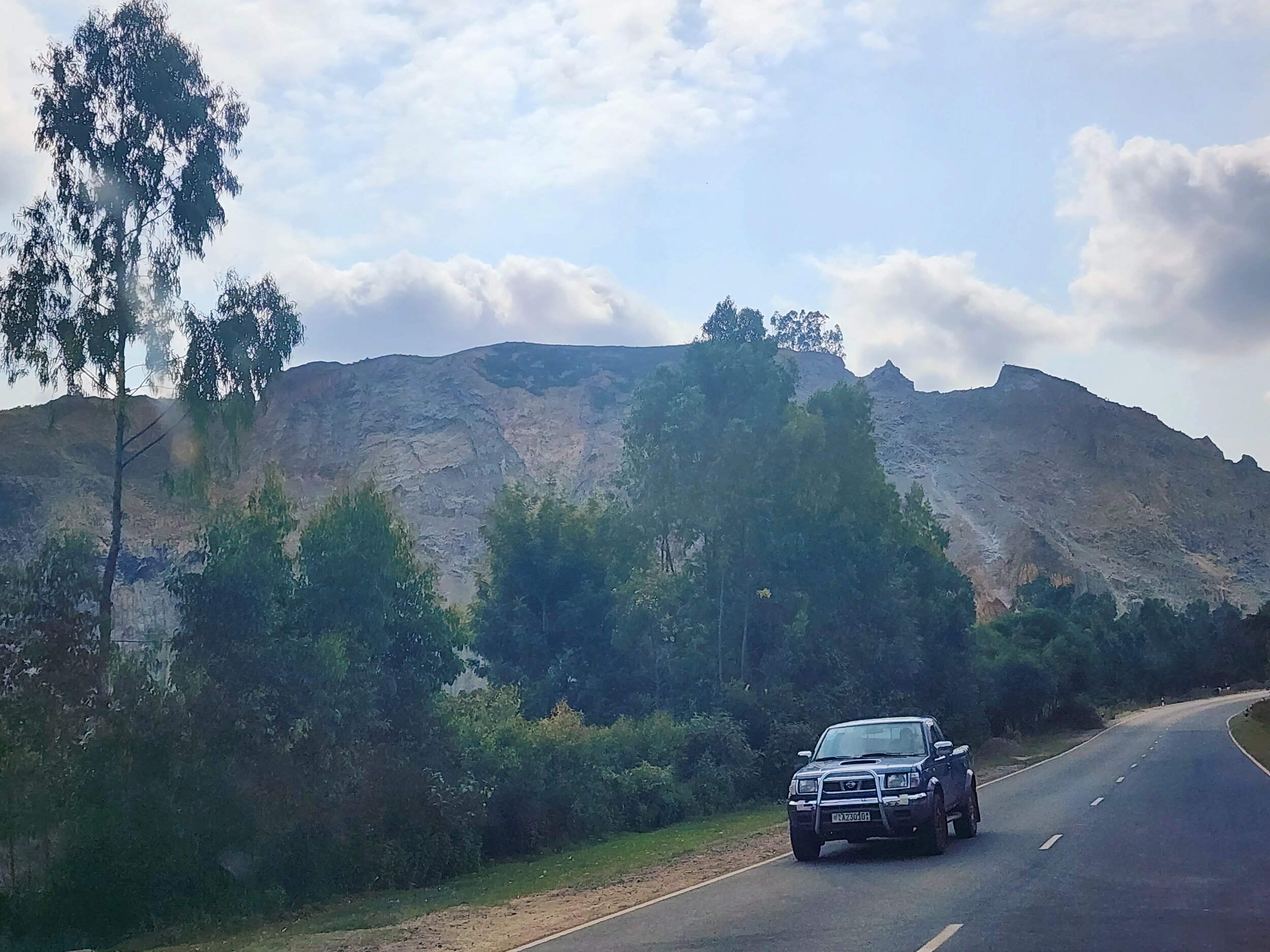 A mountain road scene with a pickup truck parked on the roadside, surrounded by trees and hills under a partly cloudy sky in Ethiopia.