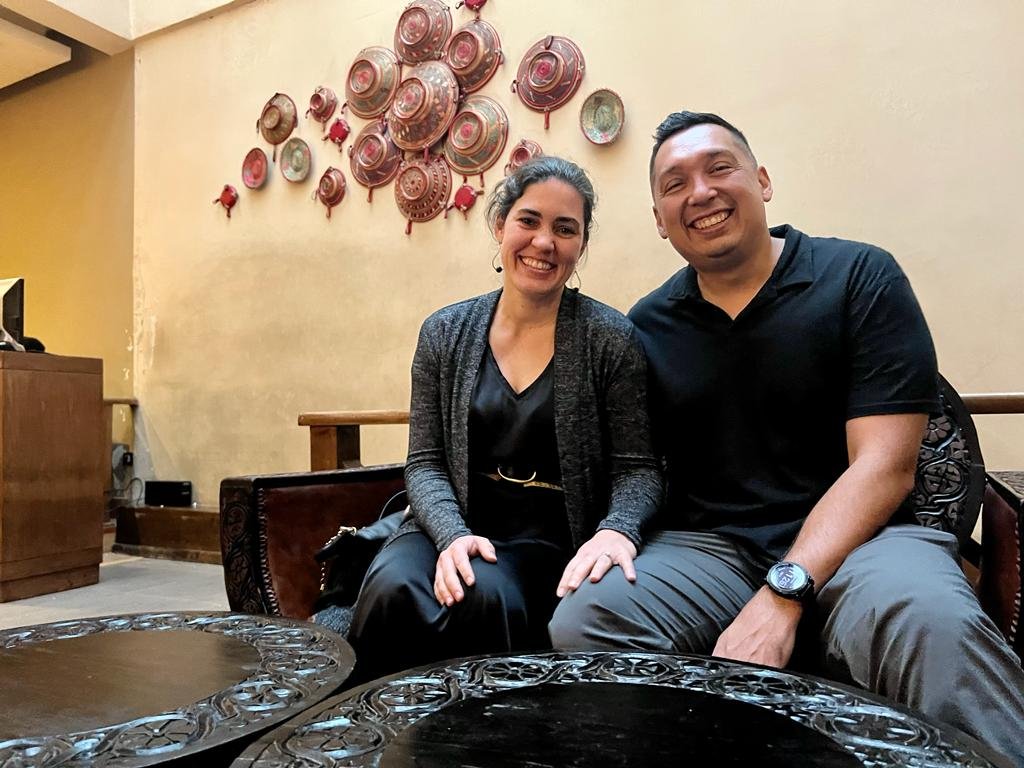 Two people smiling indoors, sitting next to each other in front of a decorative wall adorned with circular art pieces. A wooden counter is visible in the background.