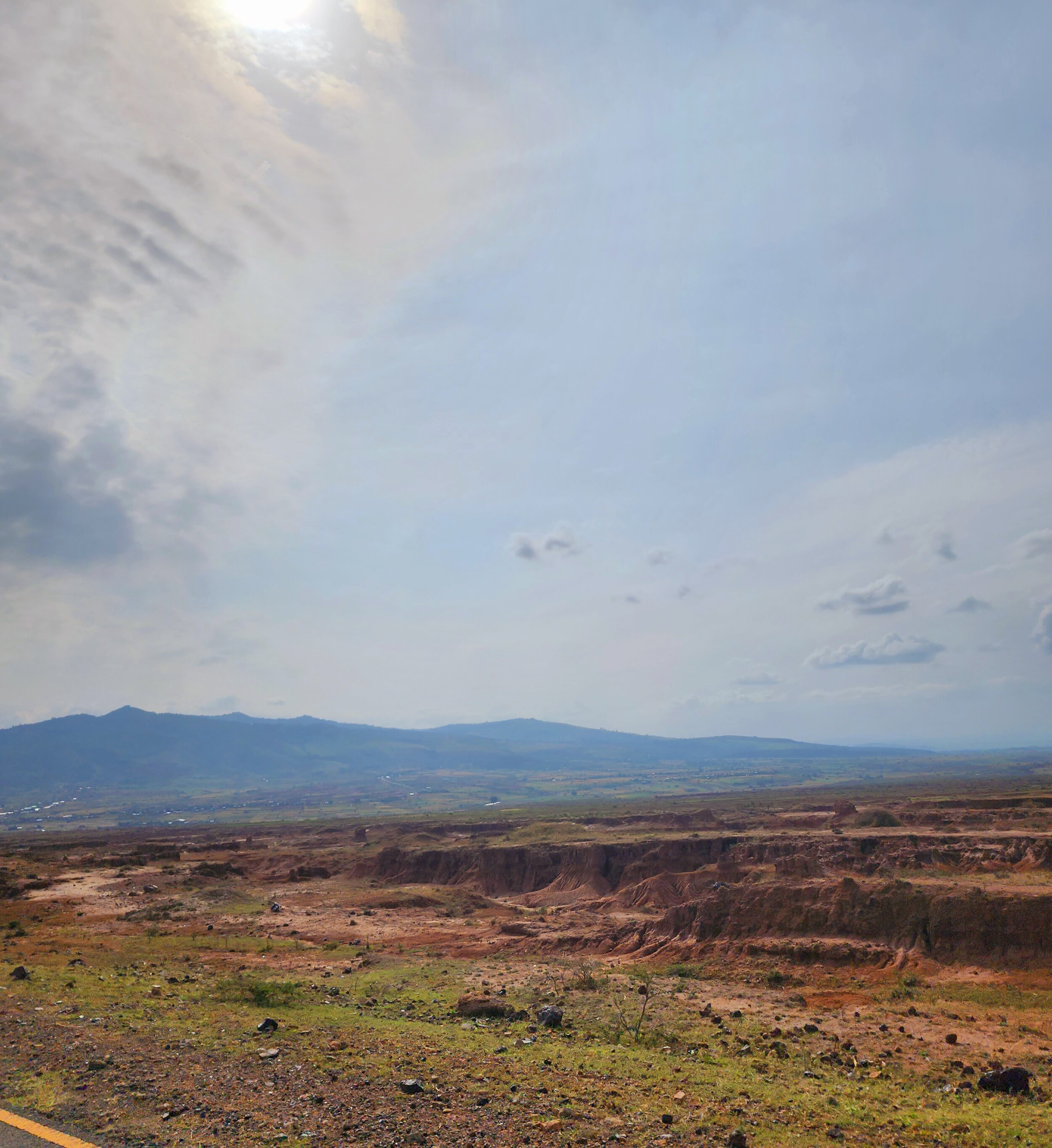 A vast, sparsely vegetated Ethiopian landscape with rolling hills under a bright, slightly cloudy sky. Sunlight peeks through the clouds, illuminating the terrain, which consists of scattered rocks and patches of grass.