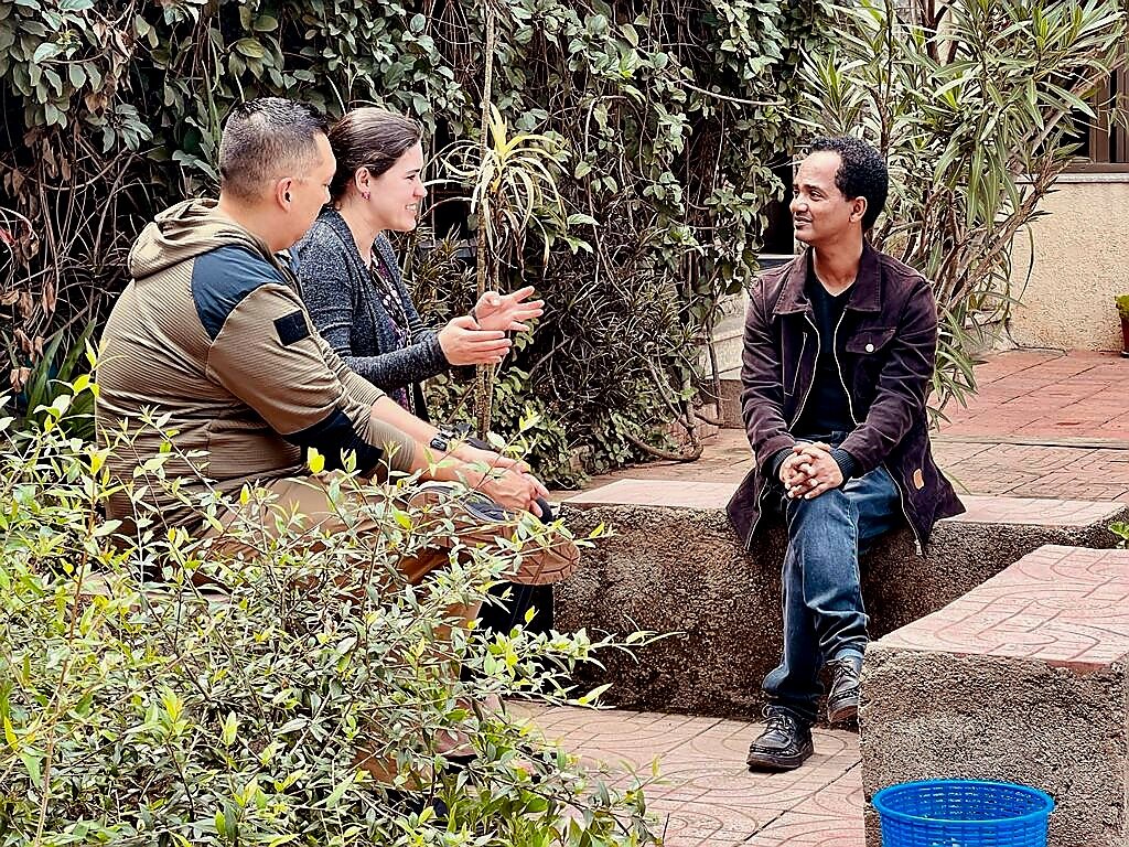 Three people having a conversation sitting outdoors; two on a low wall and one on a stone bench, surrounded by plants.