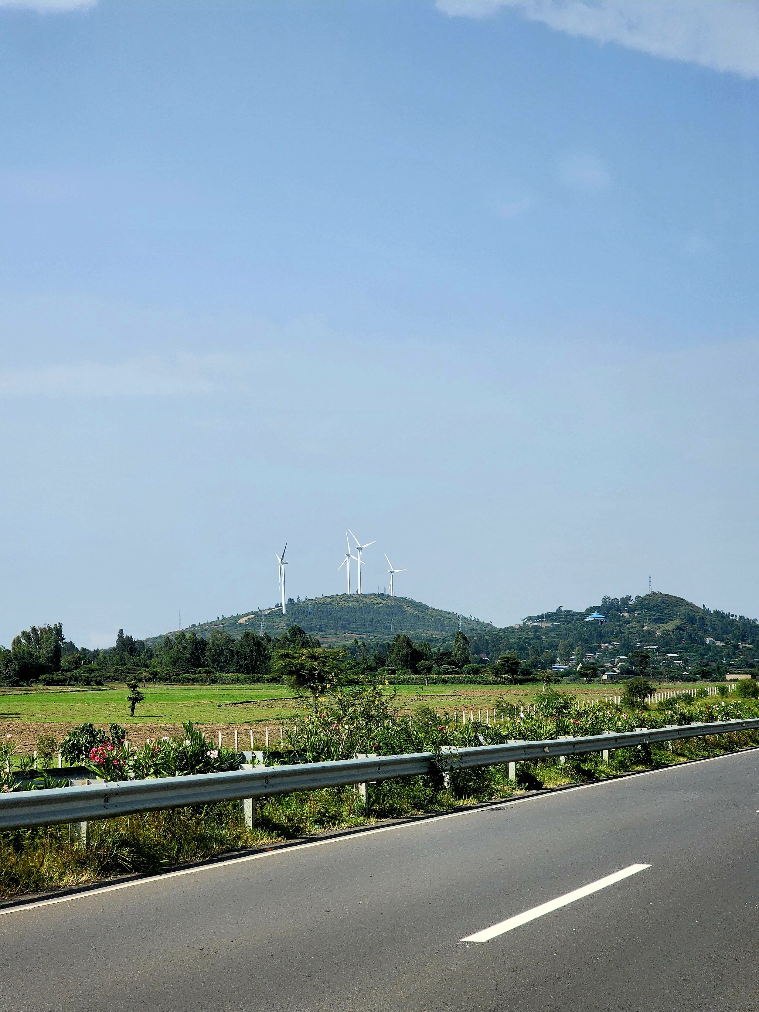 View of a road with a guardrail, open fields, hills, and wind turbines under a clear blue sky.