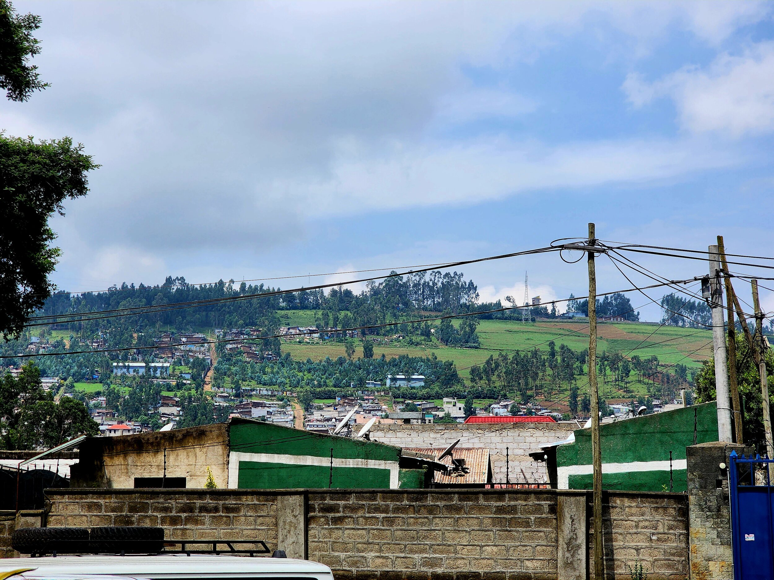 Hilly landscape with scattered houses, green fields, and trees under a cloudy sky. Power lines and poles are visible in the foreground.