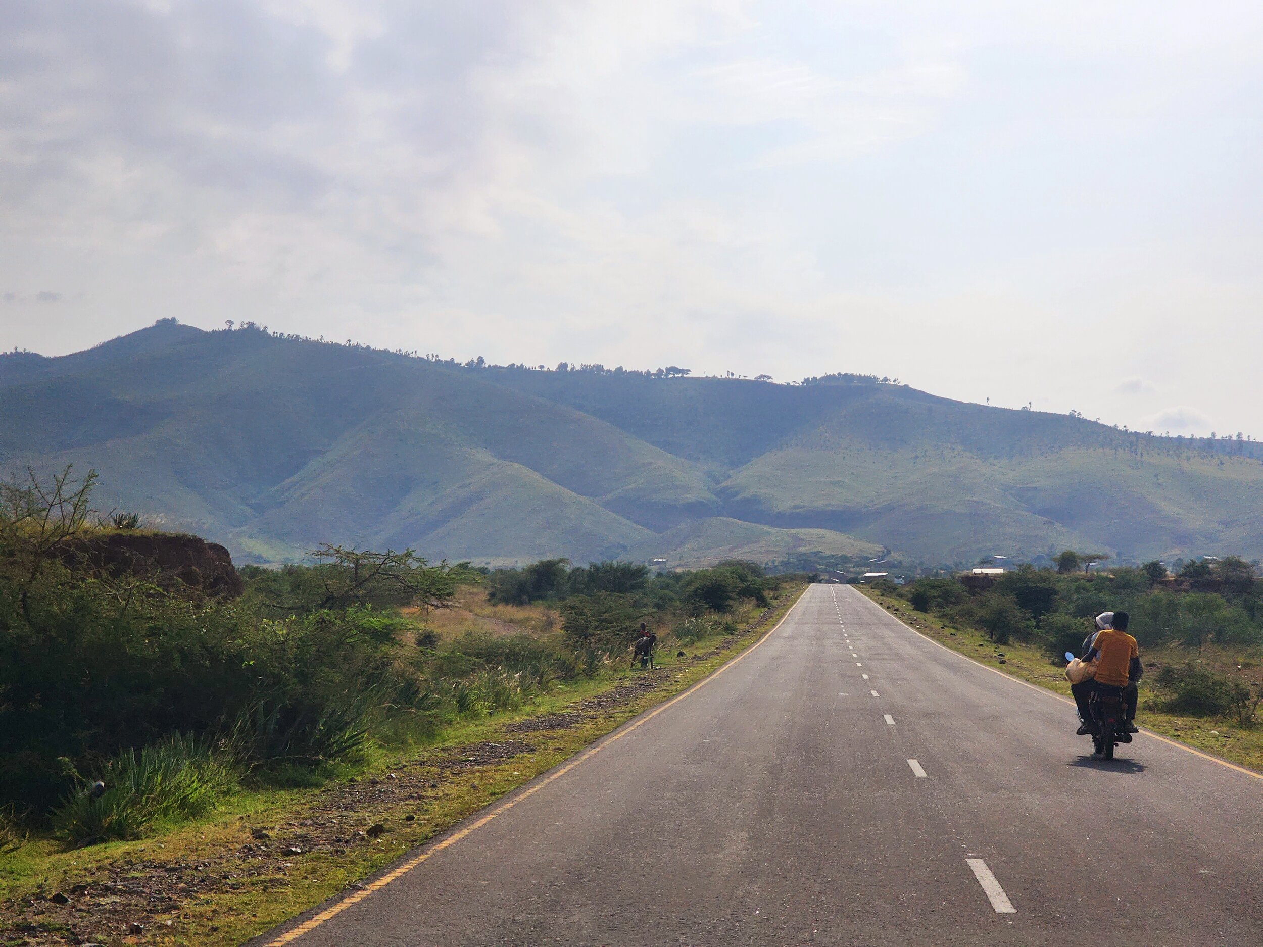 A long, straight road in Ethiopia leading towards distant green hills under cloudy skies, with two Ethiopians riding a motorcycle on the right side of the road.