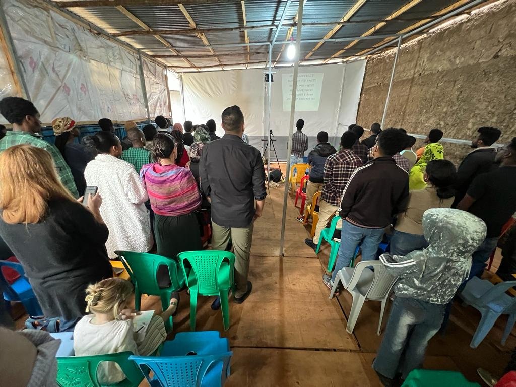 A group of people gathered in a makeshift room with plastic chairs, watching a presentation or service projected onto a screen. The audience appears attentive, and the setting is church in Sodo, Ethiopia.