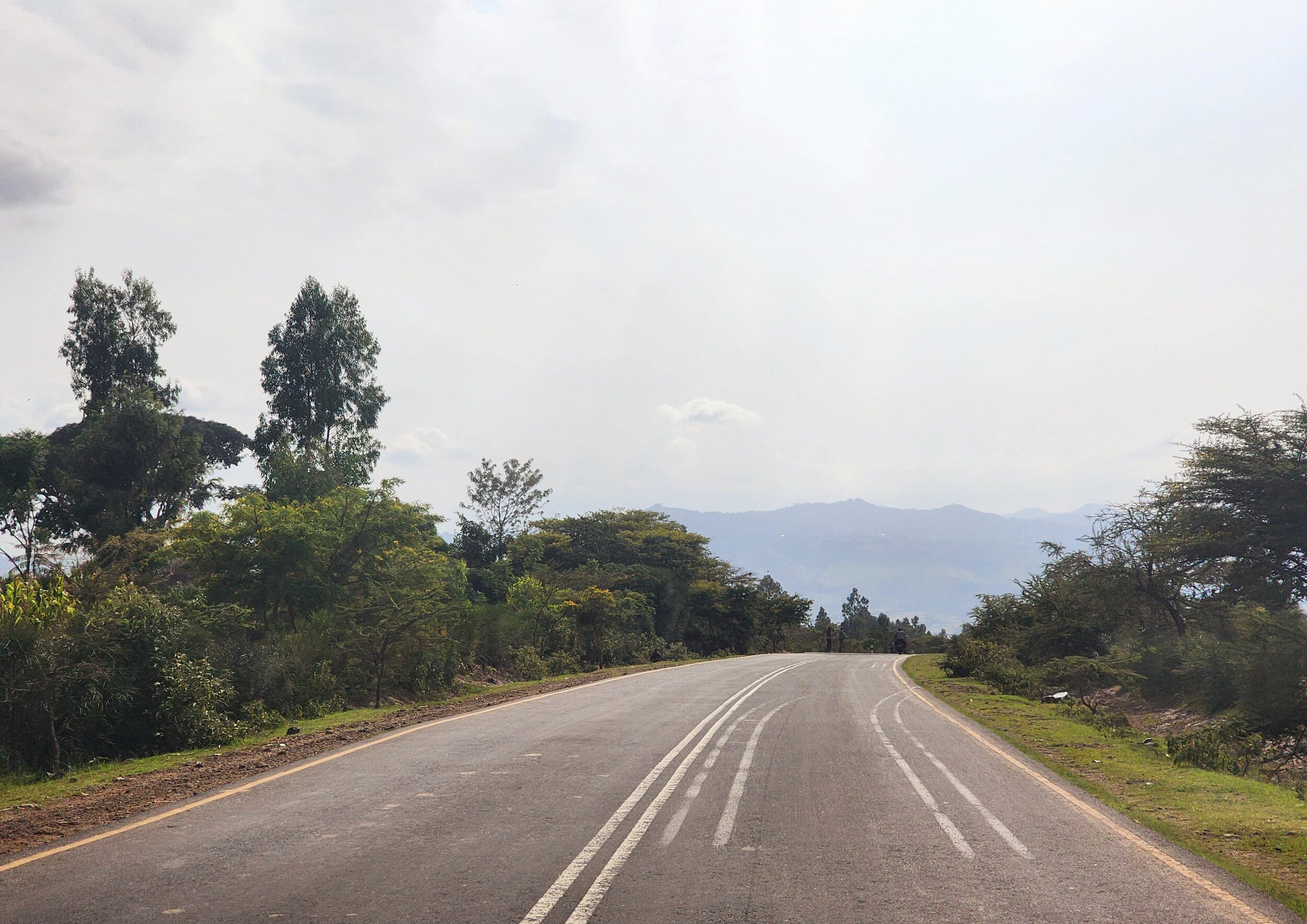 A winding road with double yellow lines surrounded by green trees and bushes, set against a background of distant hills under a cloudy sky in Ethiopia.