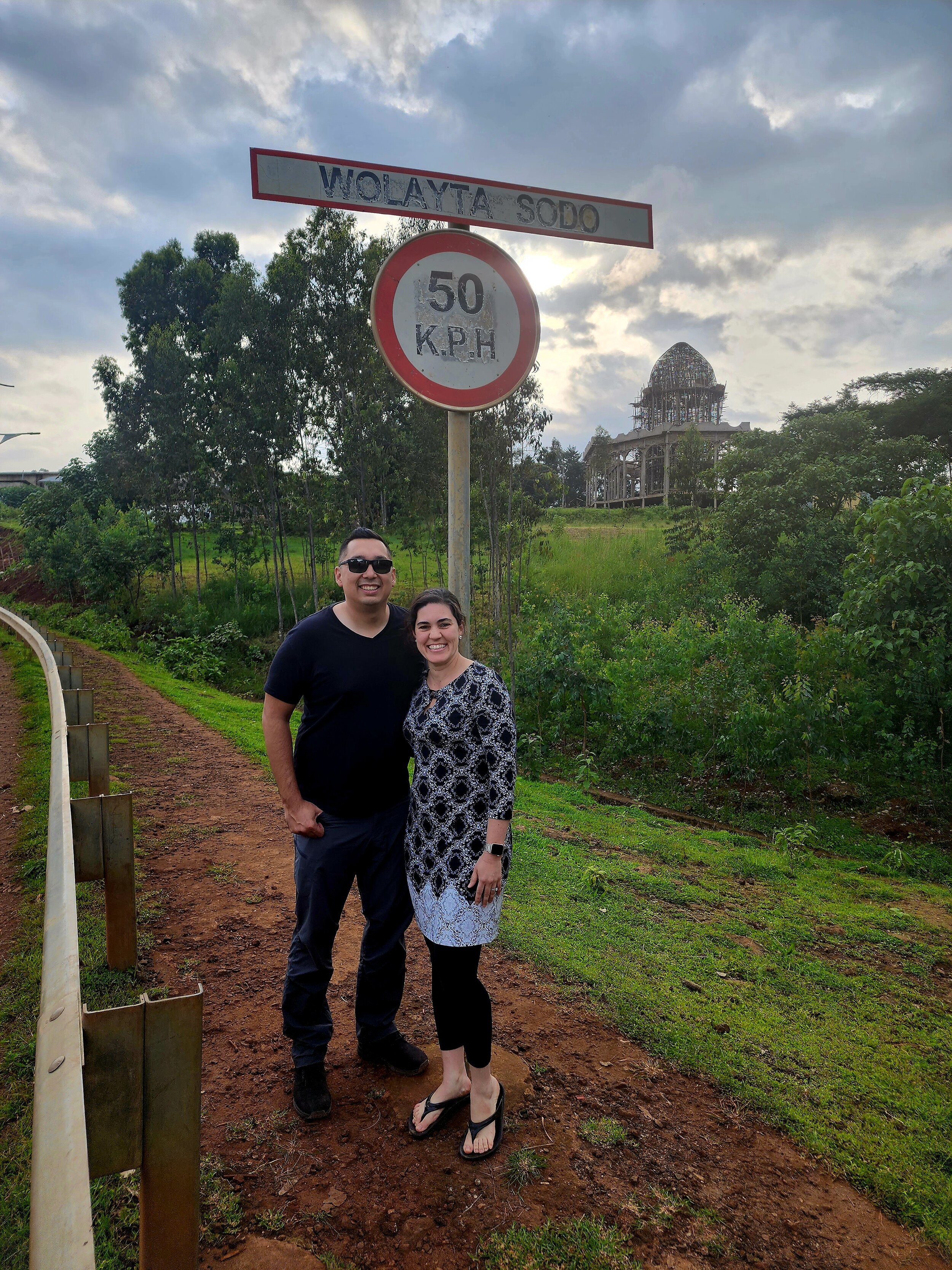 Two people standing beside a road sign indicating "WOLAYTA SODO" and "50 K.P.H" with lush greenery and a building under construction in the background, under a cloudy sky.