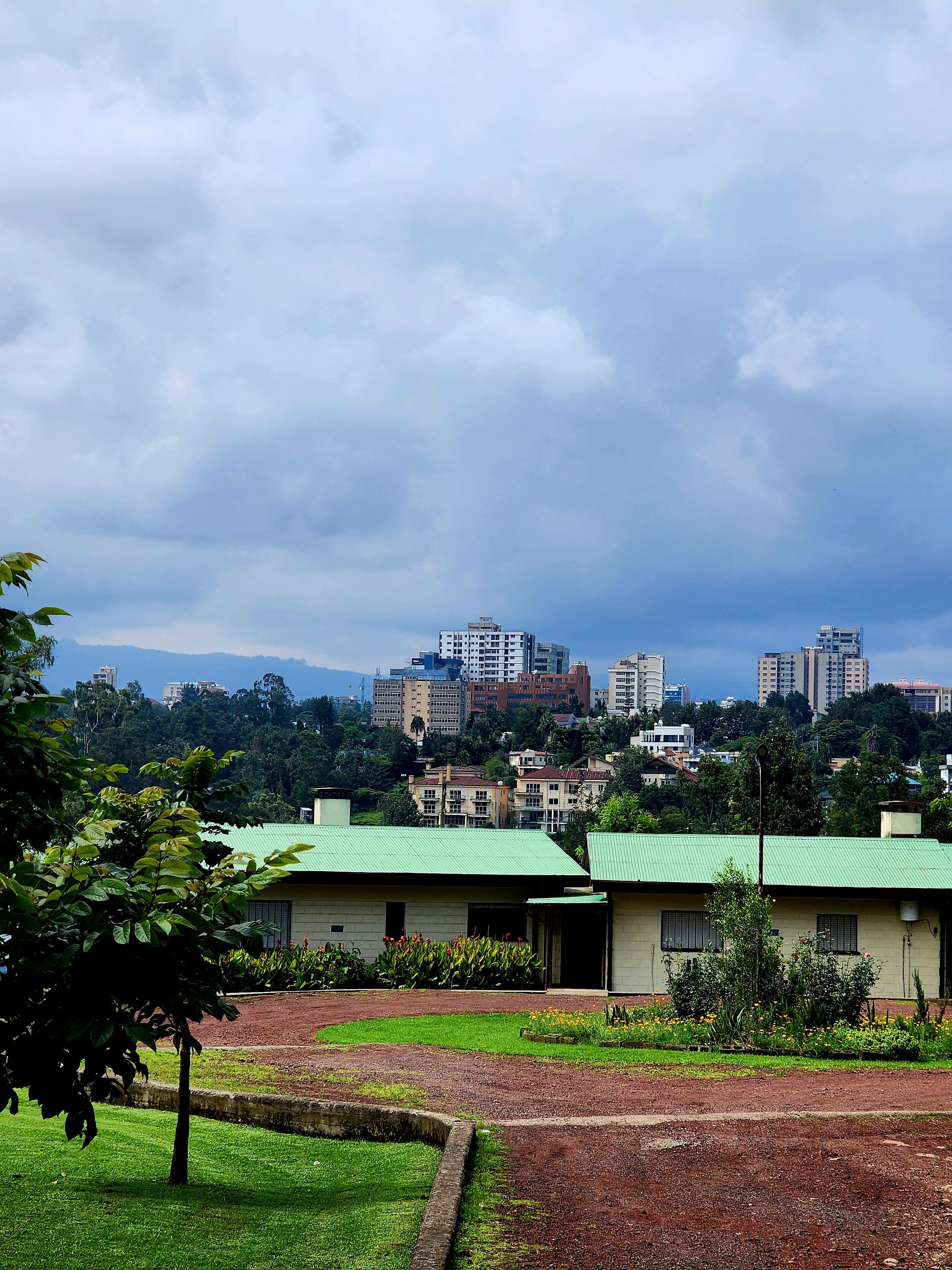 A view of green-roofed buildings with a dirt path, surrounded by trees and greenery, with a city skyline and tall buildings in the background under cloudy skies.