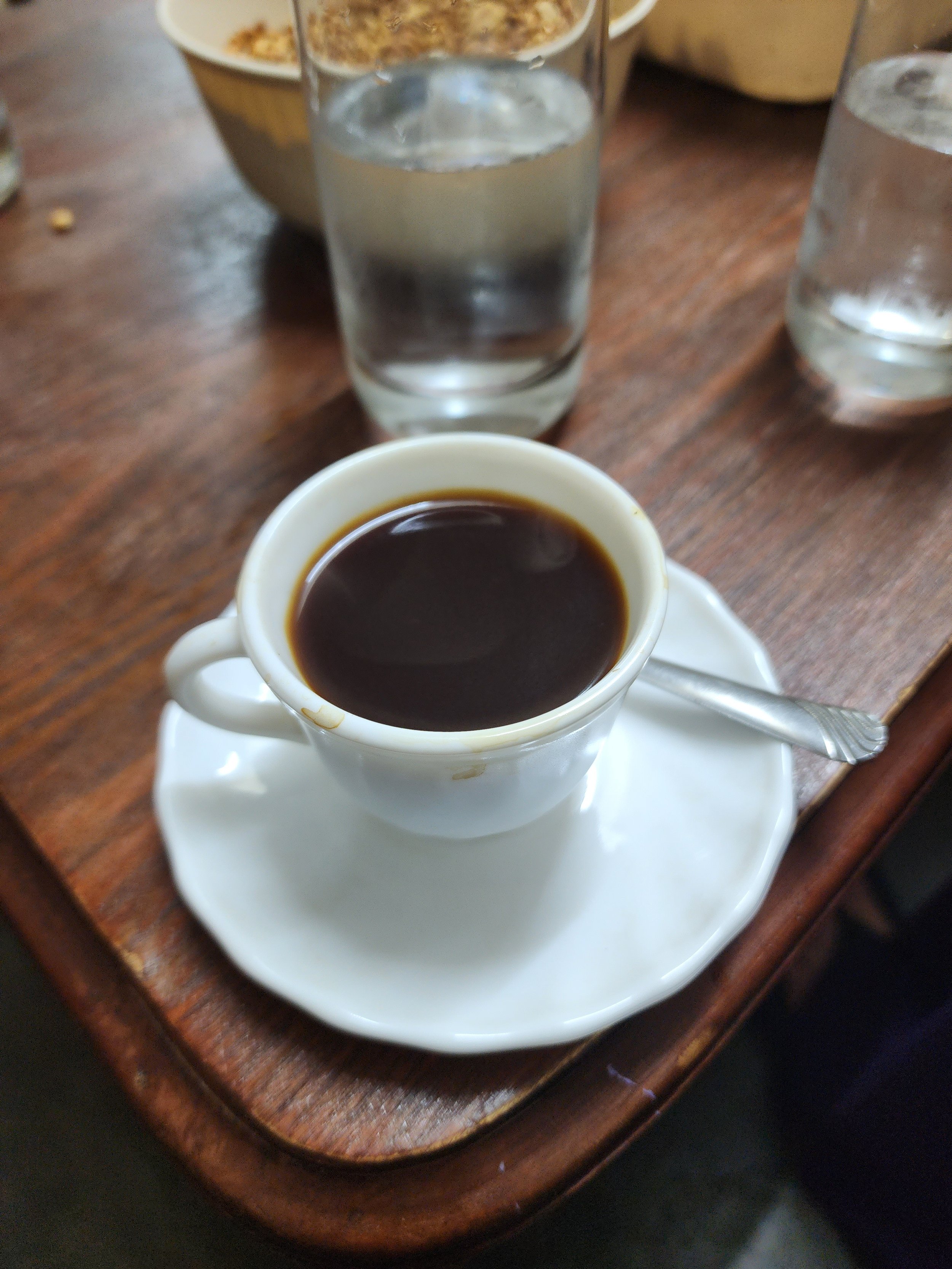 Cup of coffee with saucer and spoon on a wooden table, next to a glass of water and a bowl of food in the background.