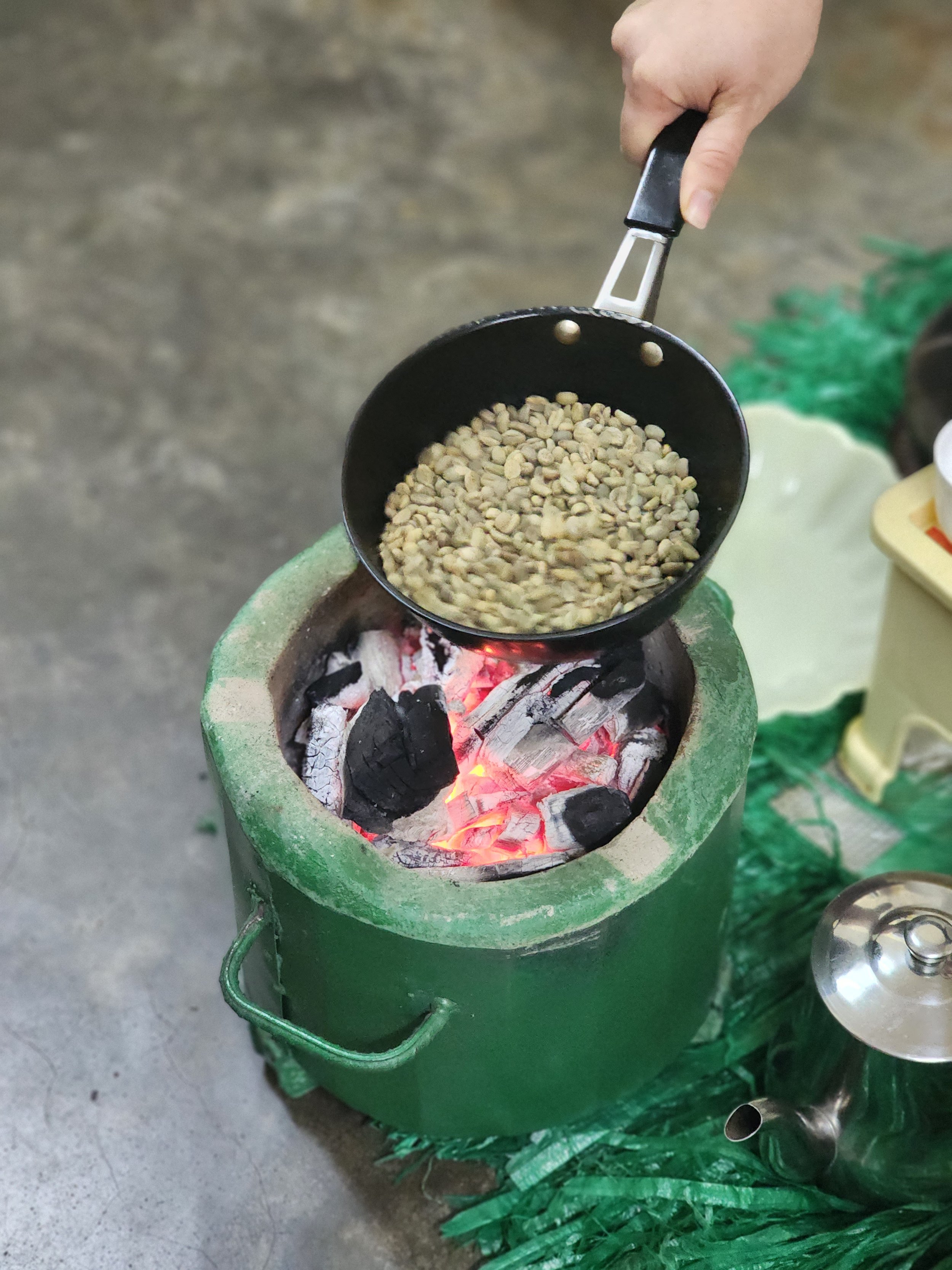 Hand holding pan with green coffee beans over a charcoal stove for roasting, next to a silver teapot on a green mat. Ethiopian coffee ceremony.