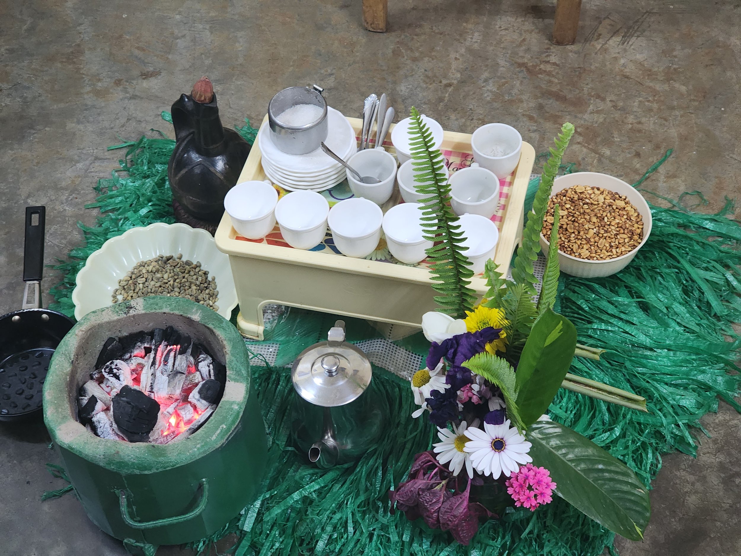 Traditional Ethiopian coffee ceremony setup with a charcoal stove, clay coffee pot, cups, roasted coffee beans, and decorative flowers on a green mat.